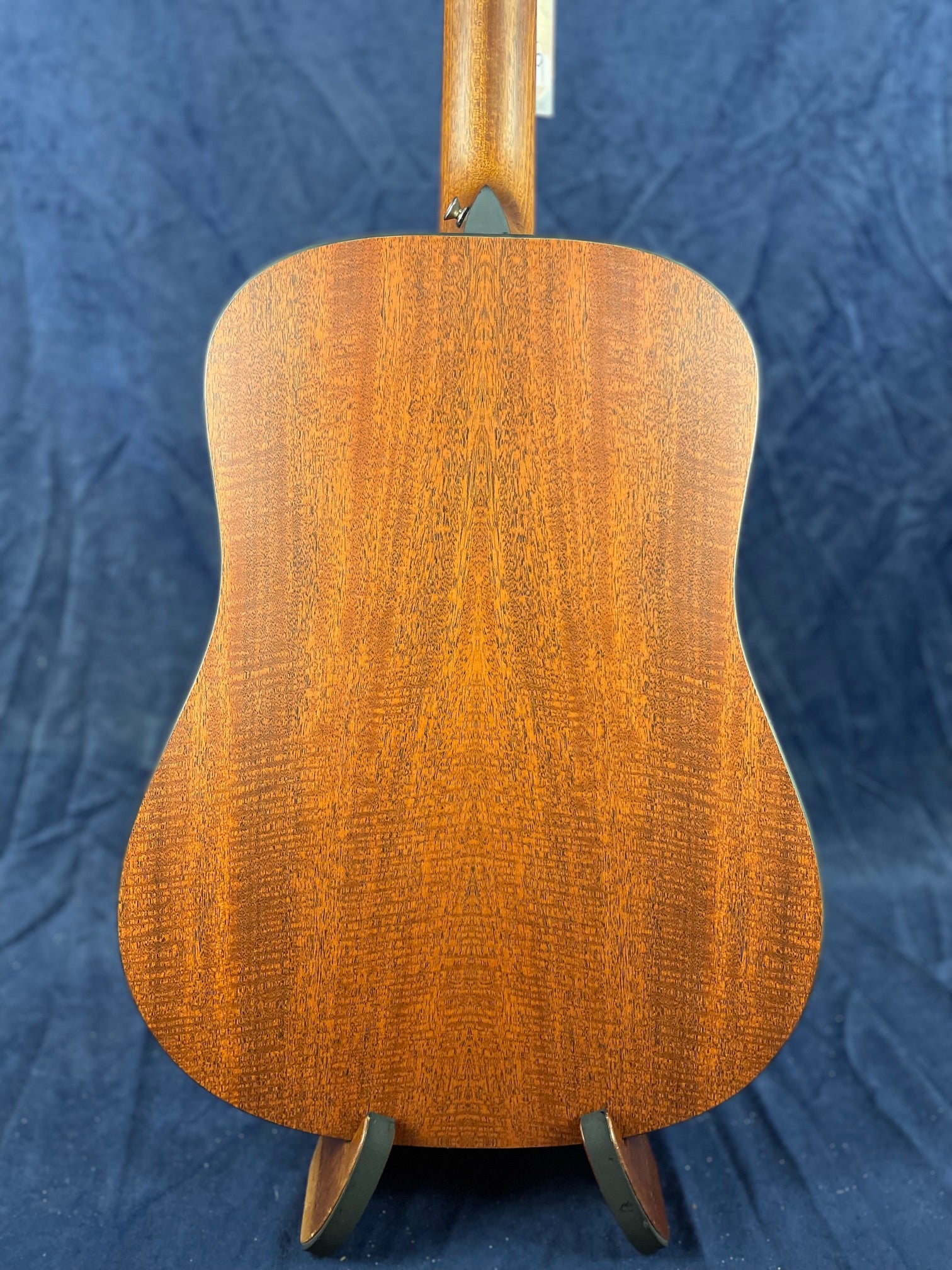 Back view of an acoustic guitar made of rich, reddish-brown wood with a straight grain pattern, shown upright against a dark blue background.