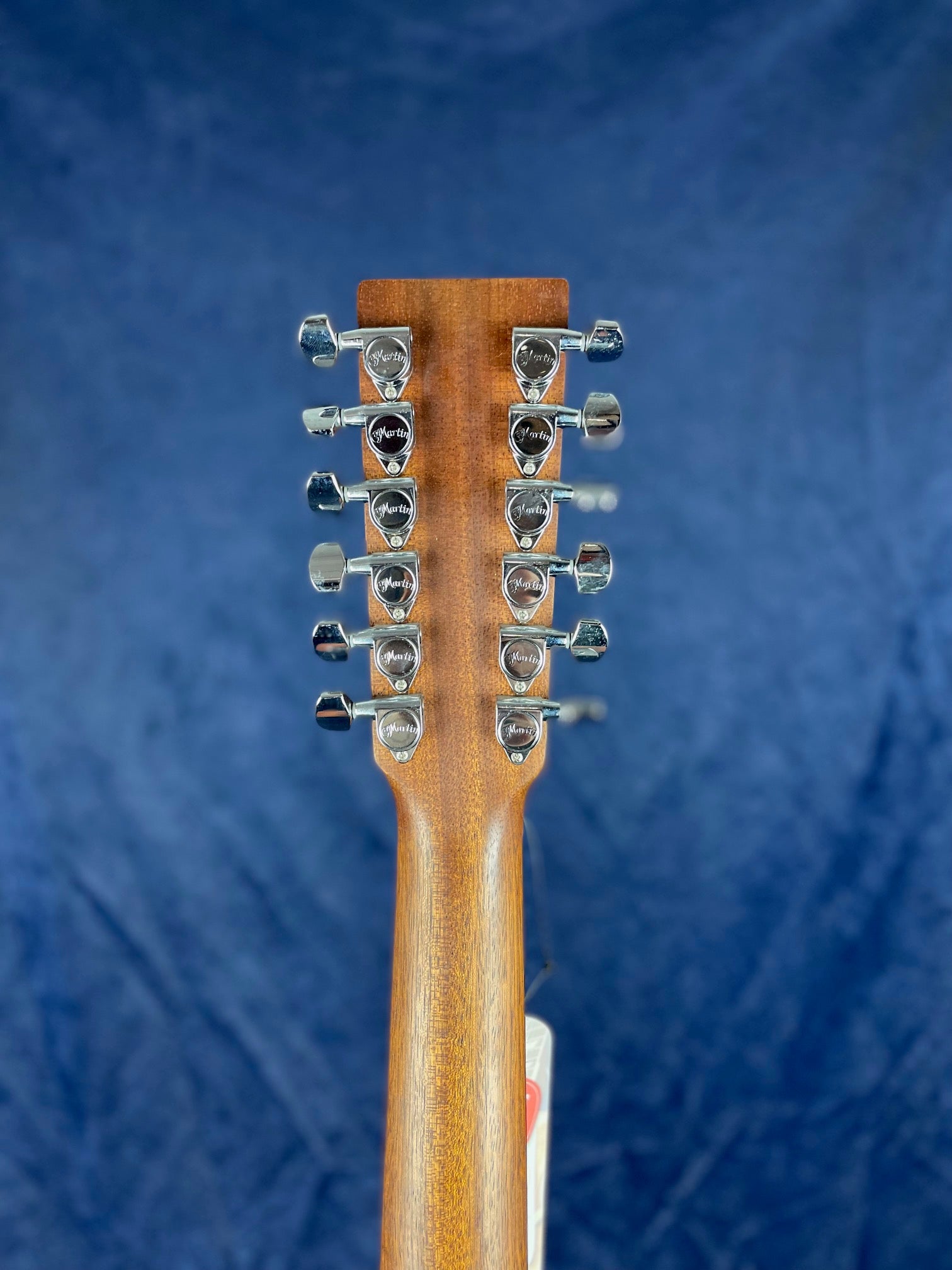 Rear view of an acoustic guitar headstock with twelve chrome tuning pegs, set against a dark blue background.