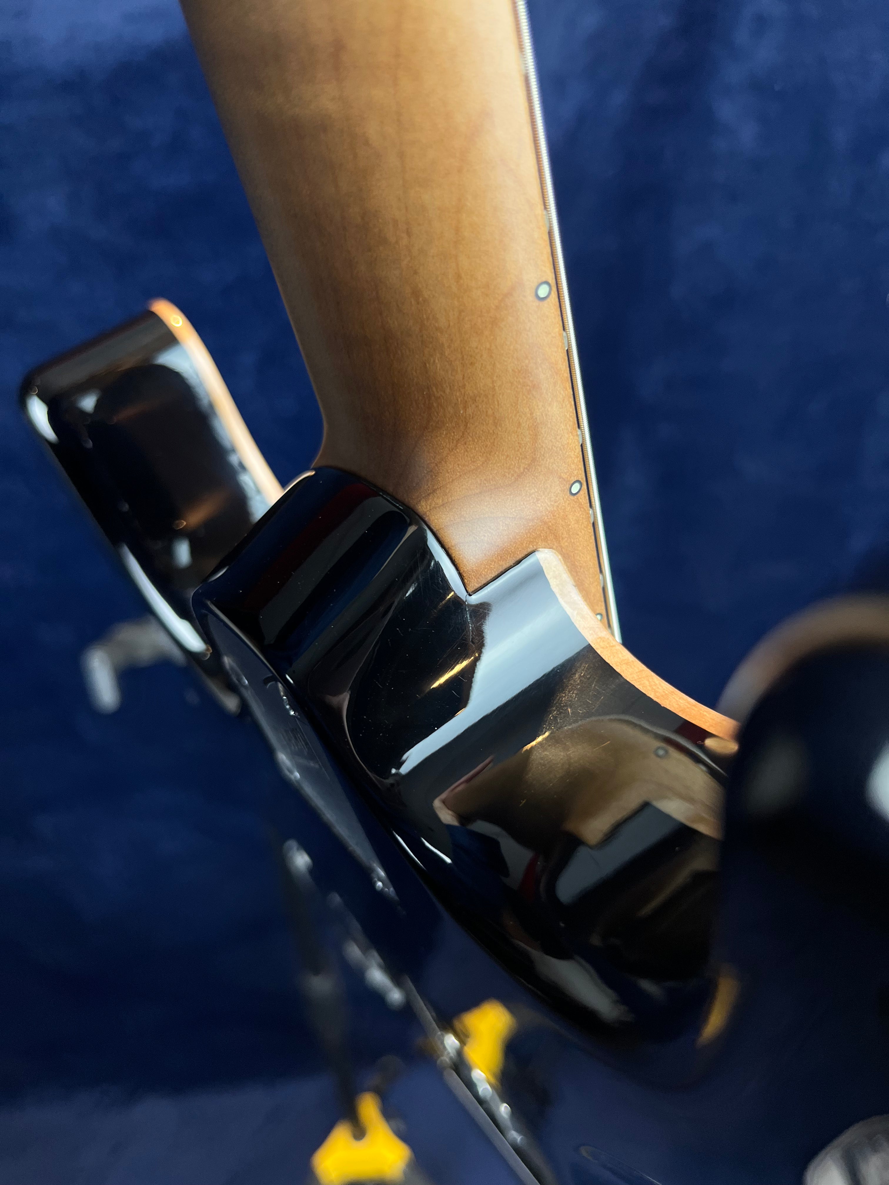 Close-up of an electric guitar’s neck joint, showing smooth wood grain transitioning to a glossy black body against a dark blue background.