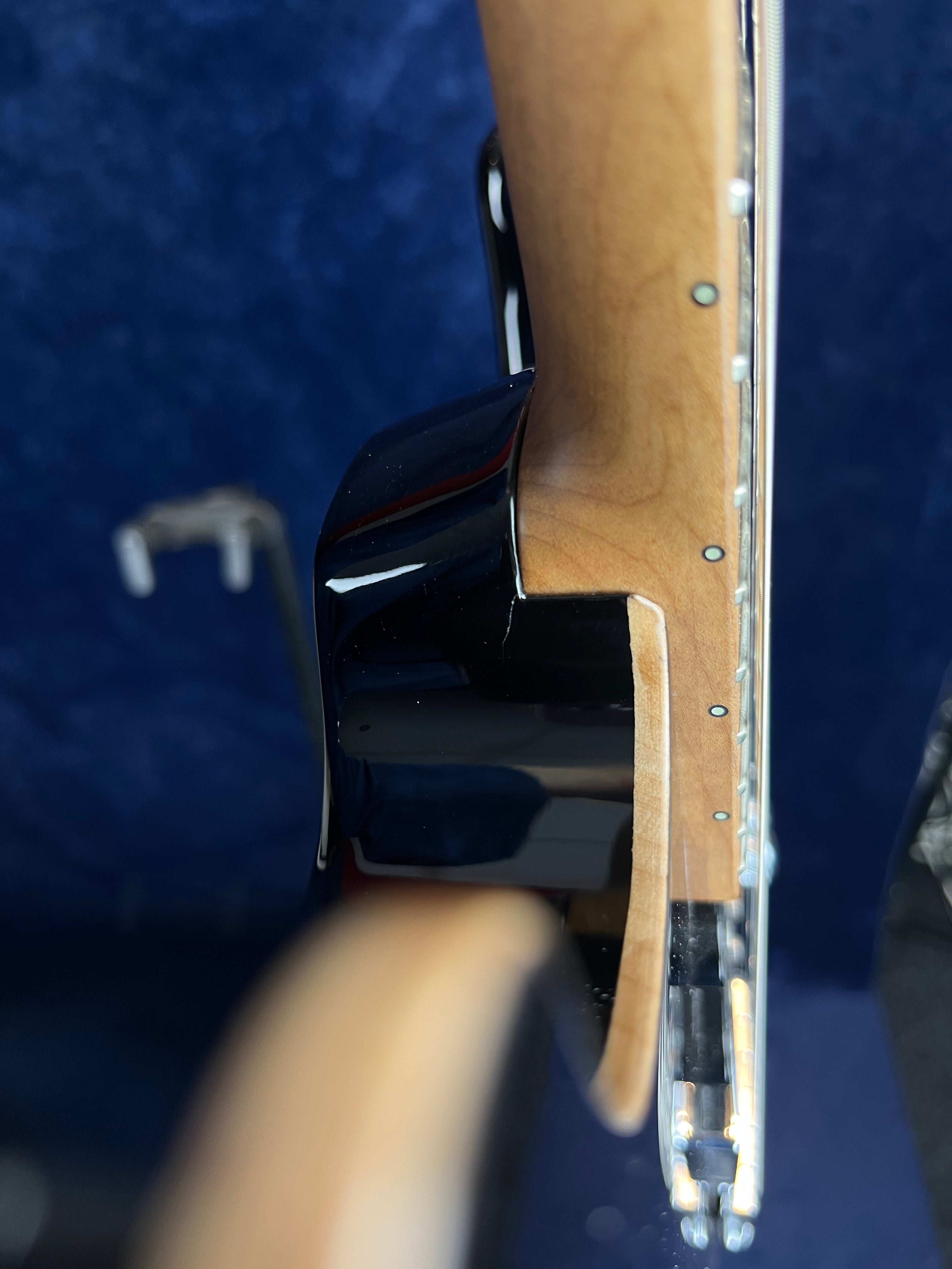 Side closeup of an electric guitar’s neck joint, showing the natural wood neck, fret markers, and glossy black body against a blue background.