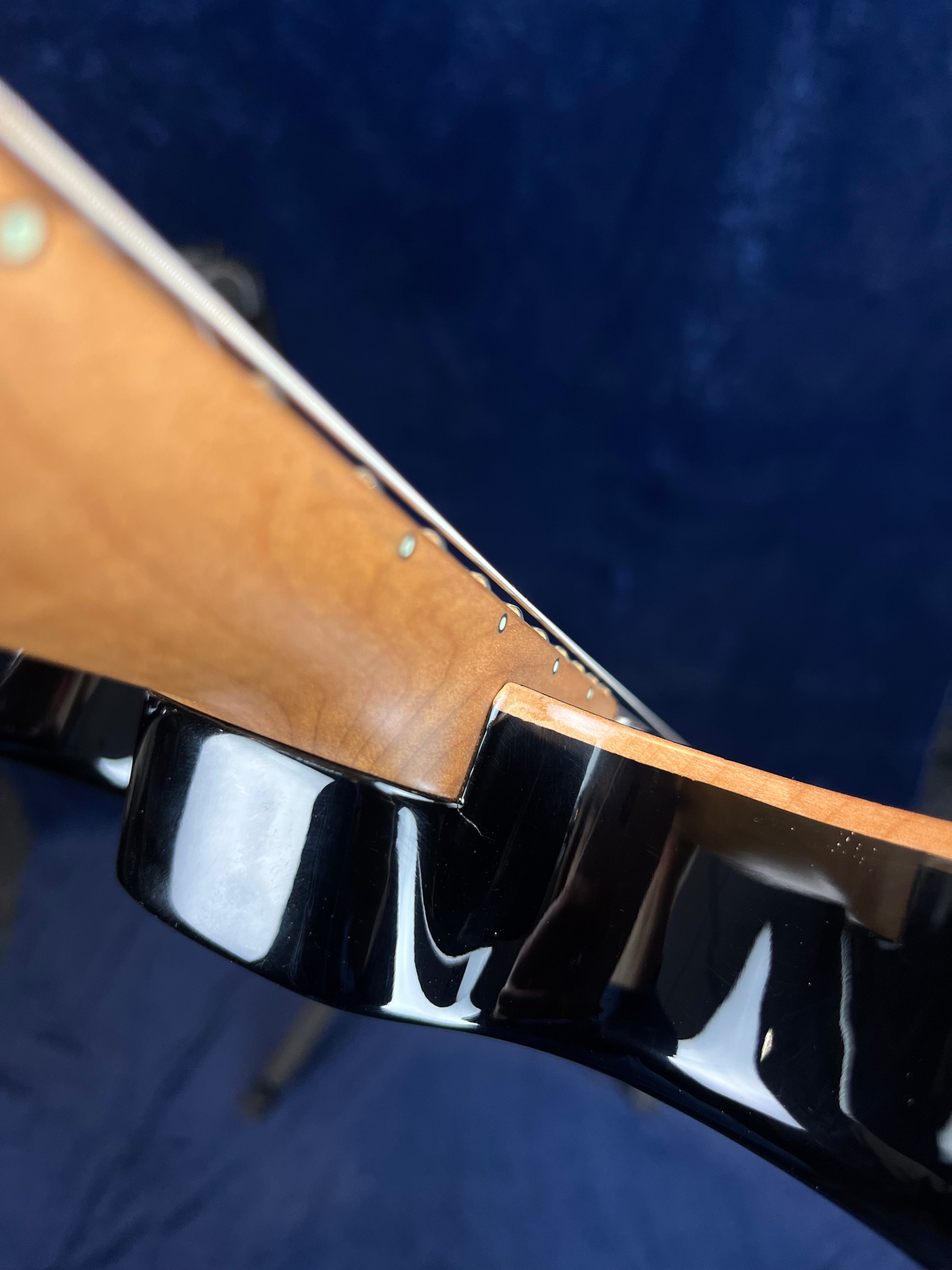 Close-up of an electric guitar neck joint with natural wood neck meeting a glossy black body, showing detailed wood grain and finish transition.