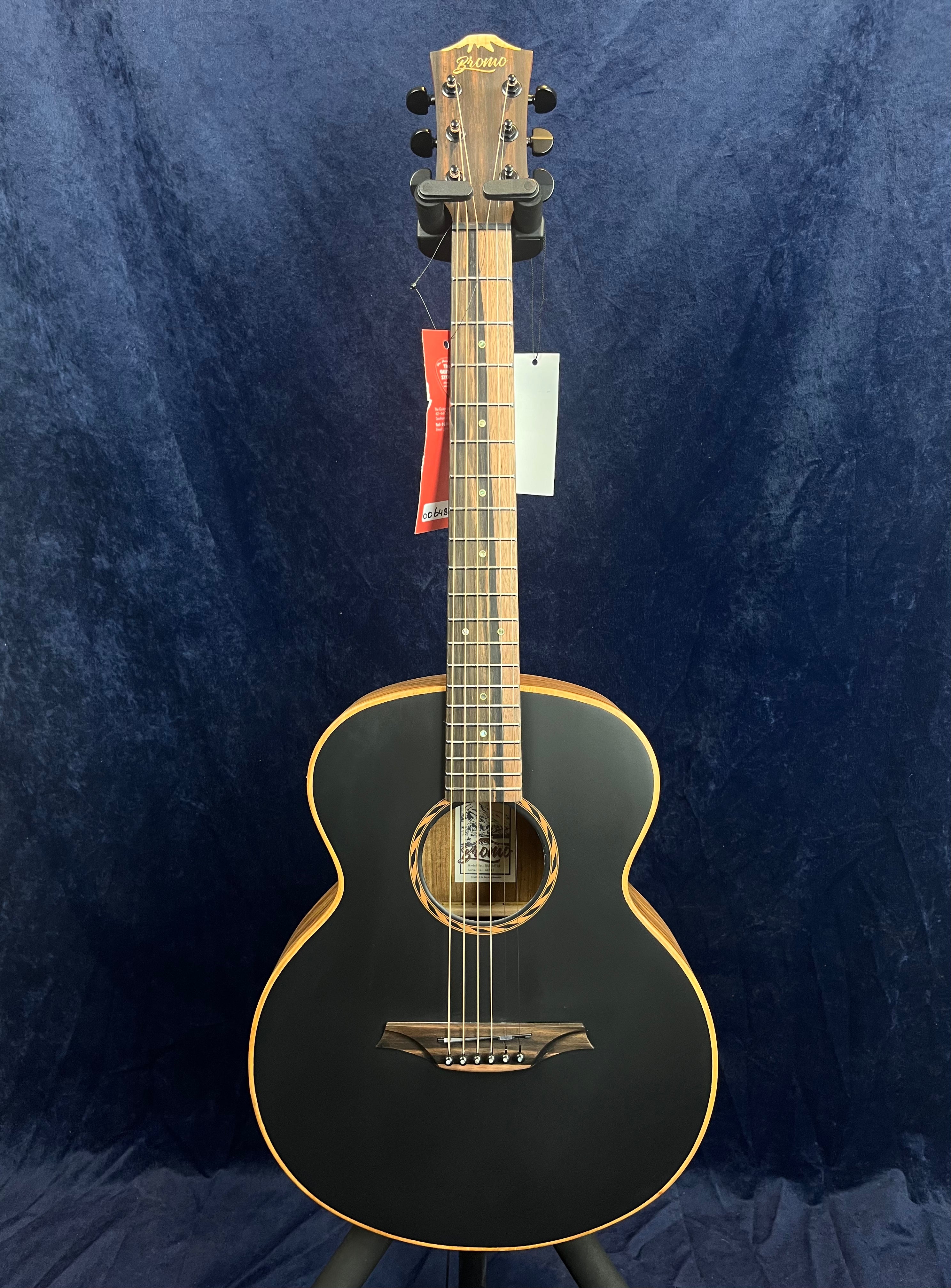 Guitare acoustique noire à contour beige, manche en bois clair, tête assortie, six cordes, rosace décorée, étiquettes suspendues au manche.