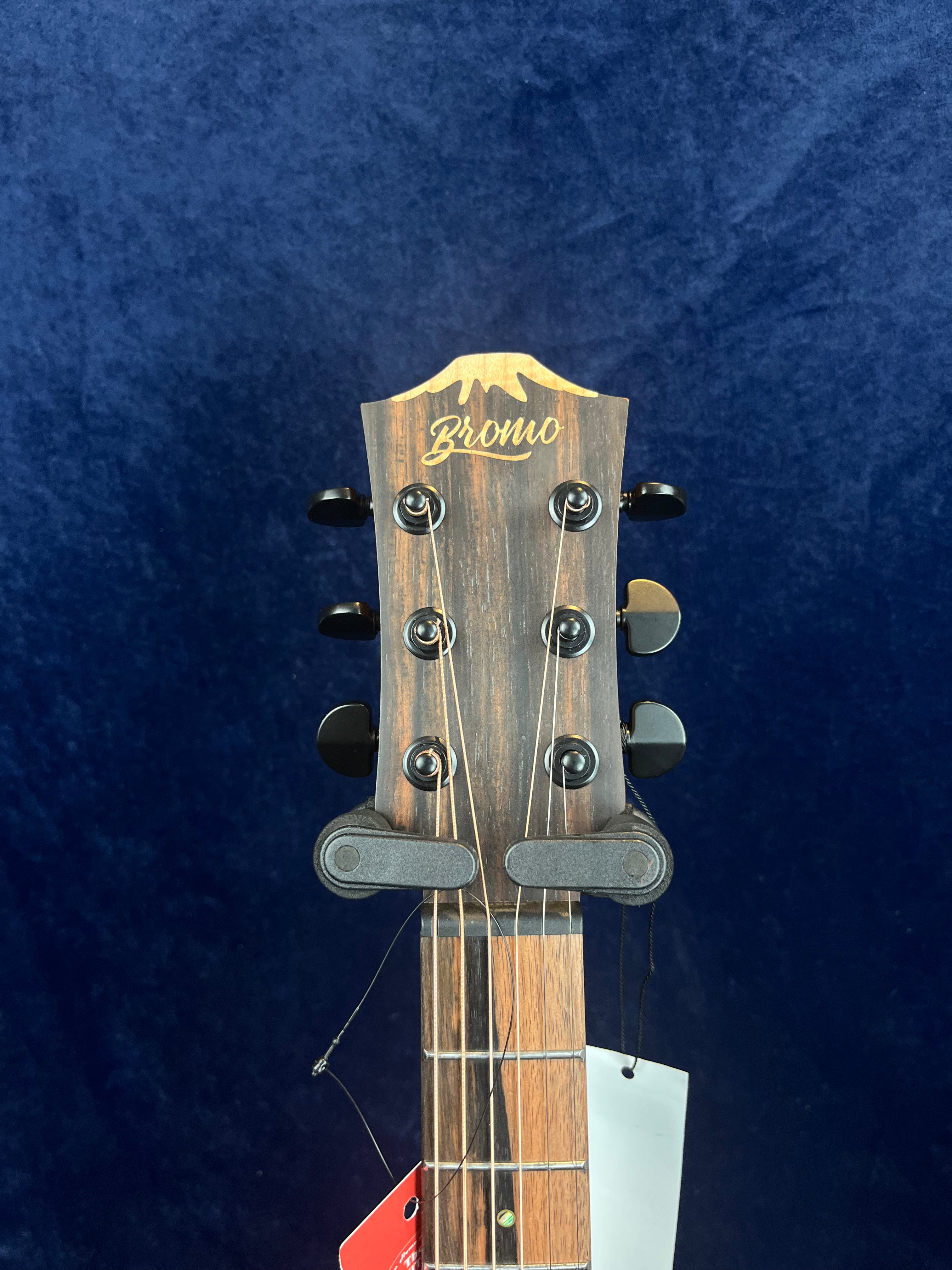 Close-up of a Bromo acoustic guitar headstock with six black tuning pegs, metallic strings, and wood grain visible against a dark blue background.