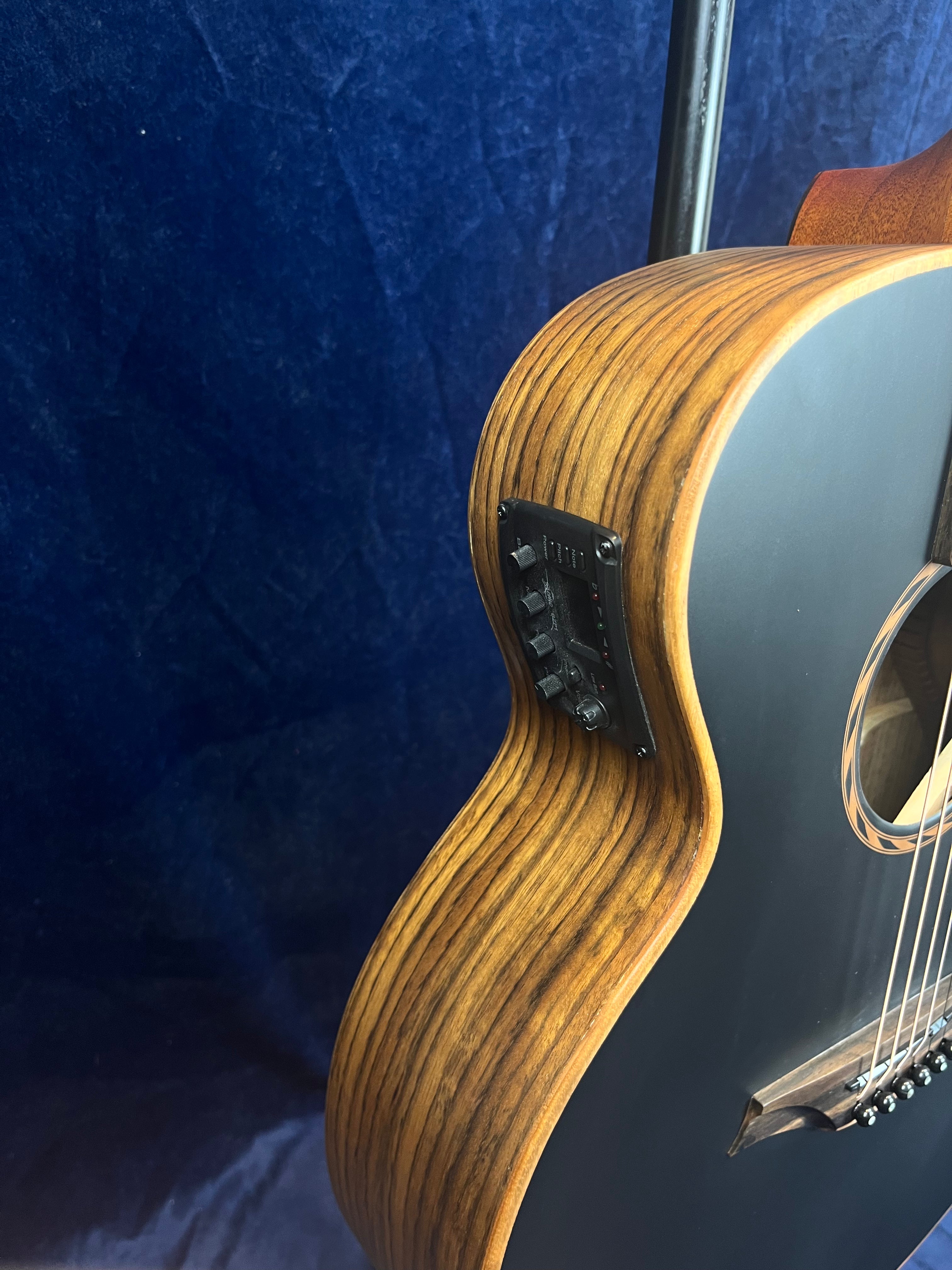 Acoustic guitar with a wood grain body, matte black top, and an electronic control panel on the side for built-in pickup or preamp.