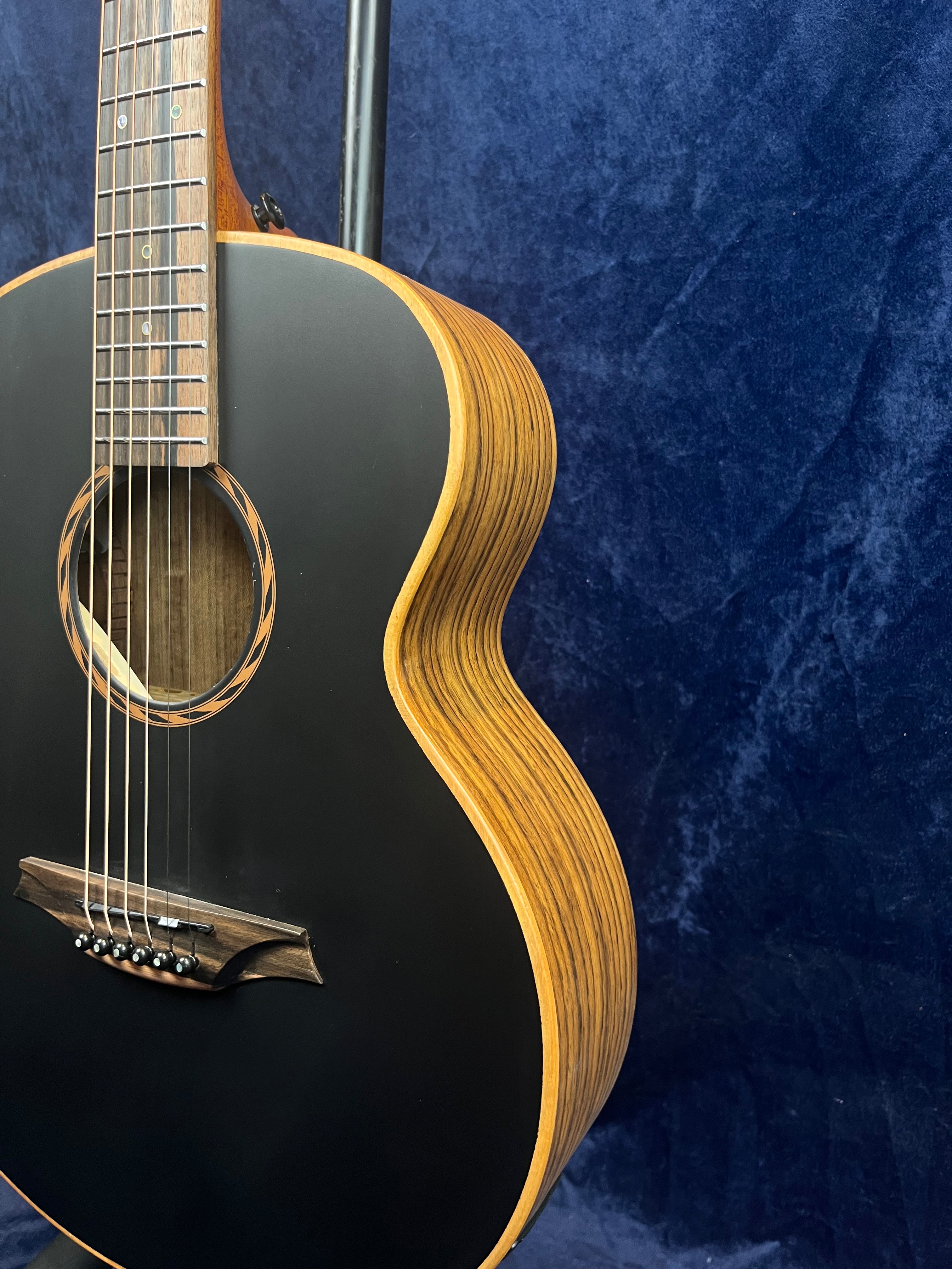 Acoustic guitar with a matte black top, natural wood sides, six strings, and a distinctive striped wood grain pattern on the body edges.