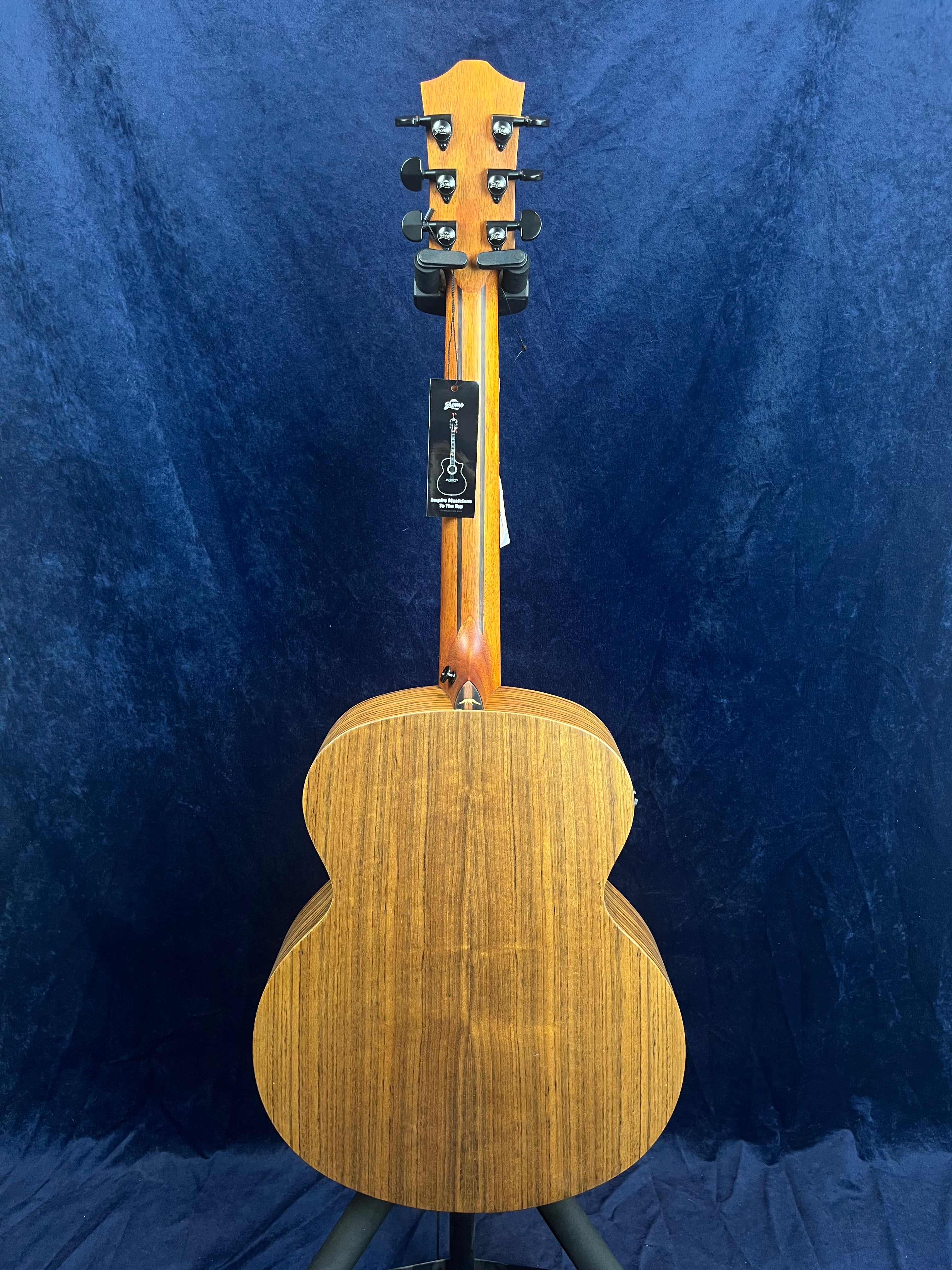 The back view of an acoustic guitar shows a natural wood finish with visible grain, black tuning pegs, and a tag hanging from the headstock.