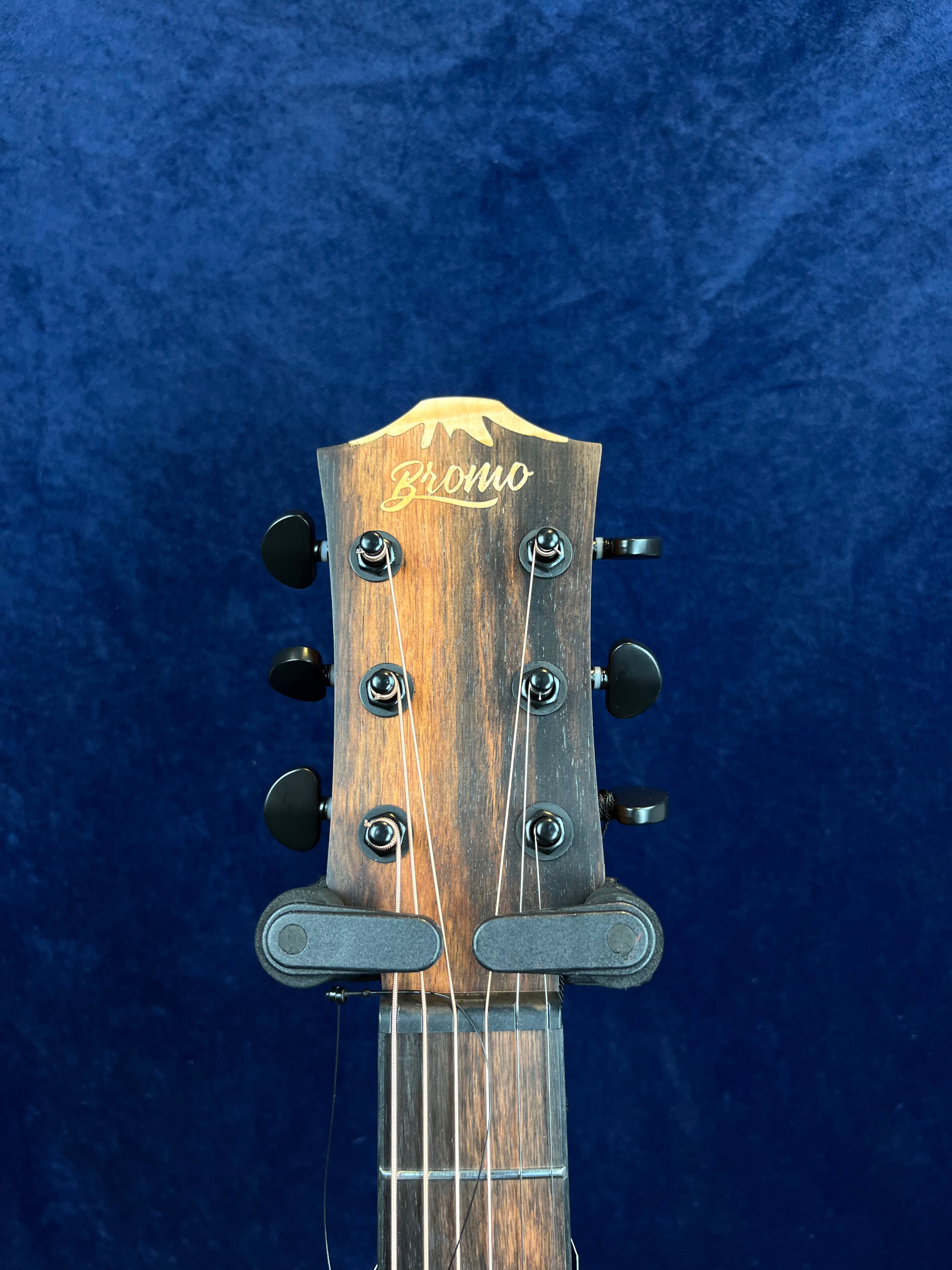 Close-up of a Bromo acoustic guitar headstock with six black tuning pegs against a dark blue background.