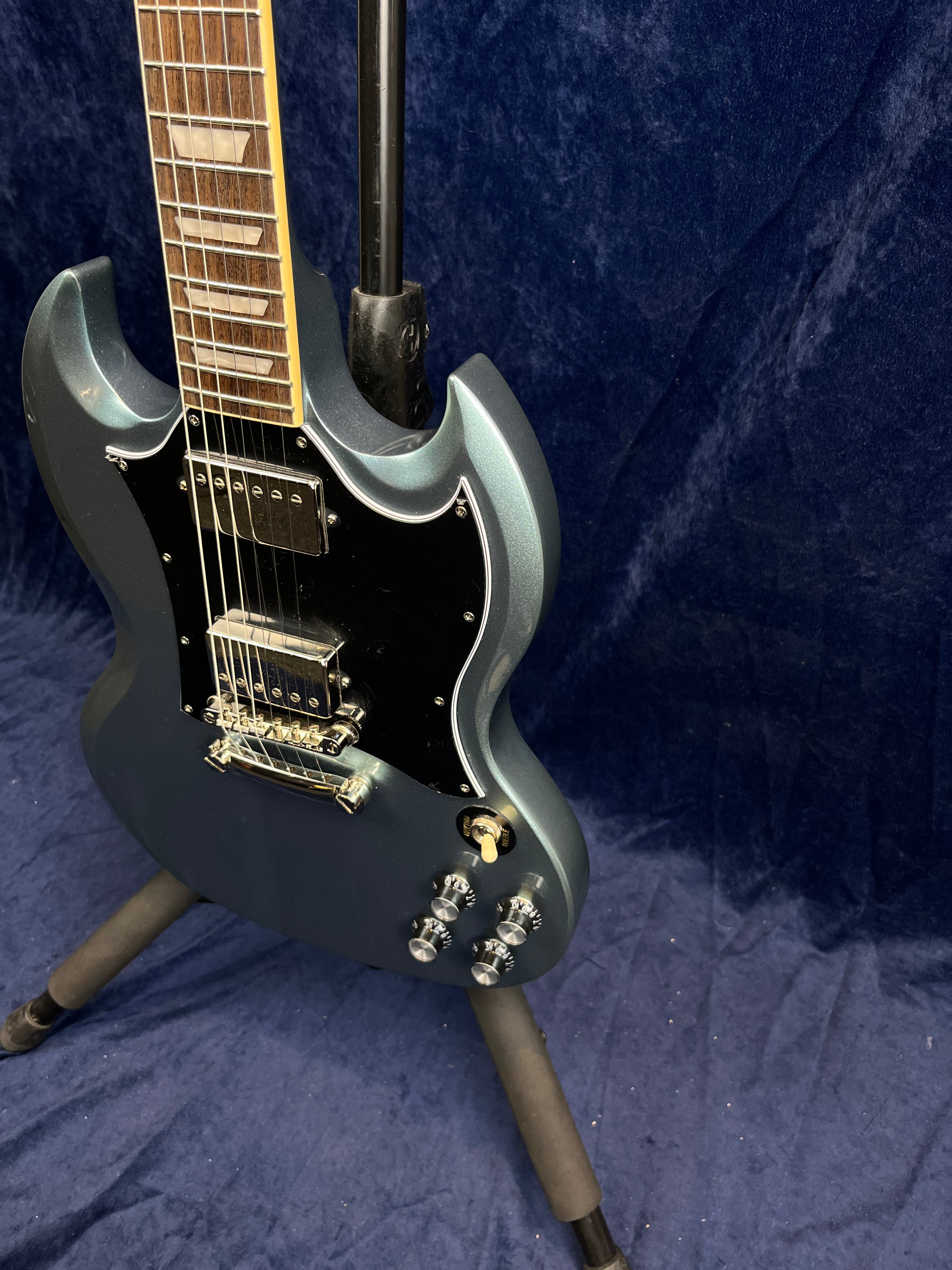 Metallic blue electric guitar with double cutaway body, two humbucker pickups, four control knobs, and a black pickguard, shown on a stand.