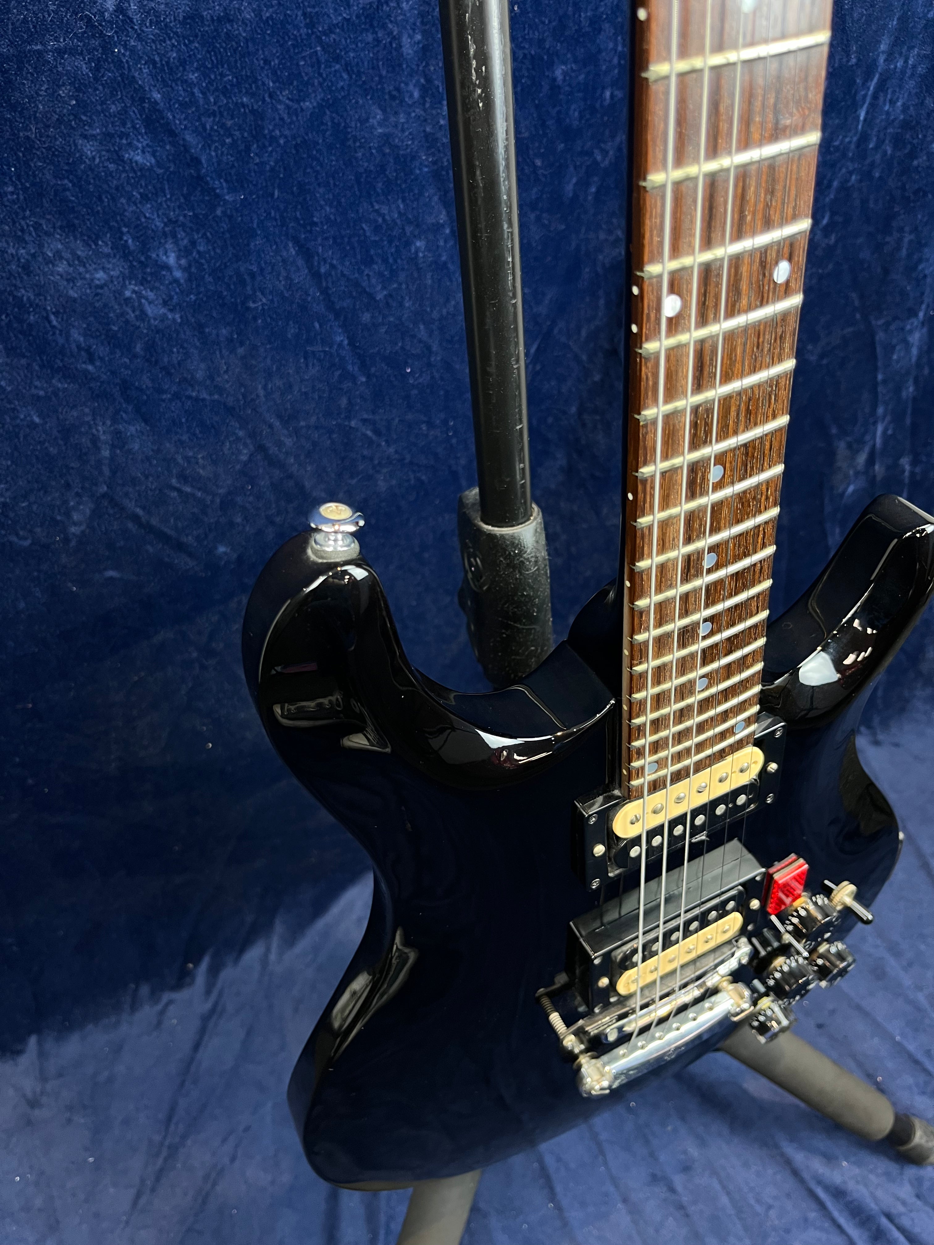 Glossy black electric guitar on a stand, showing a rosewood fretboard, dual humbucker pickups, and a tremolo bridge with adjustment knobs.