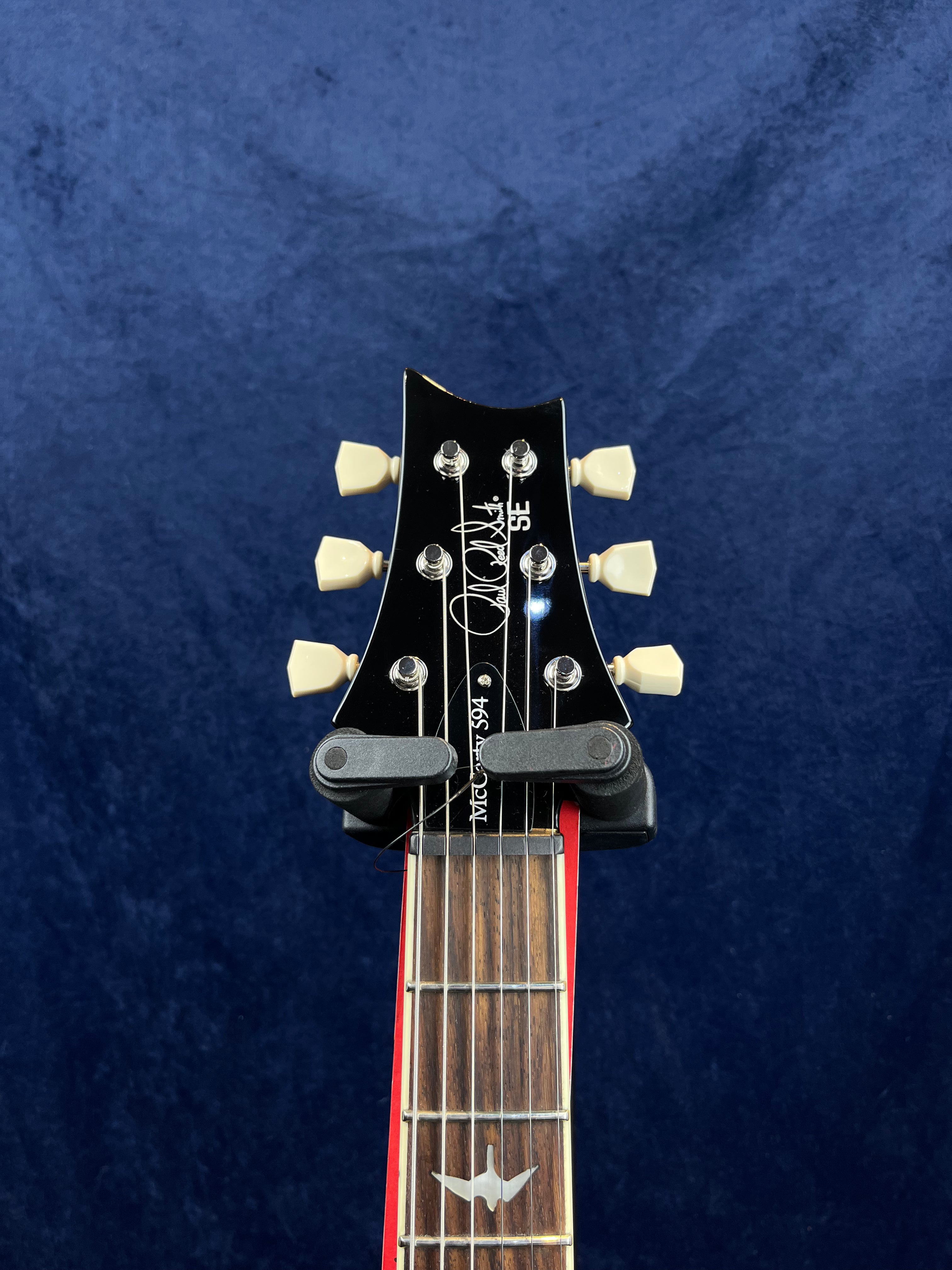 PRS SE electric guitar headstock with six cream tuners, capo attached, and a bird inlay on the rosewood fretboard against a dark blue background.