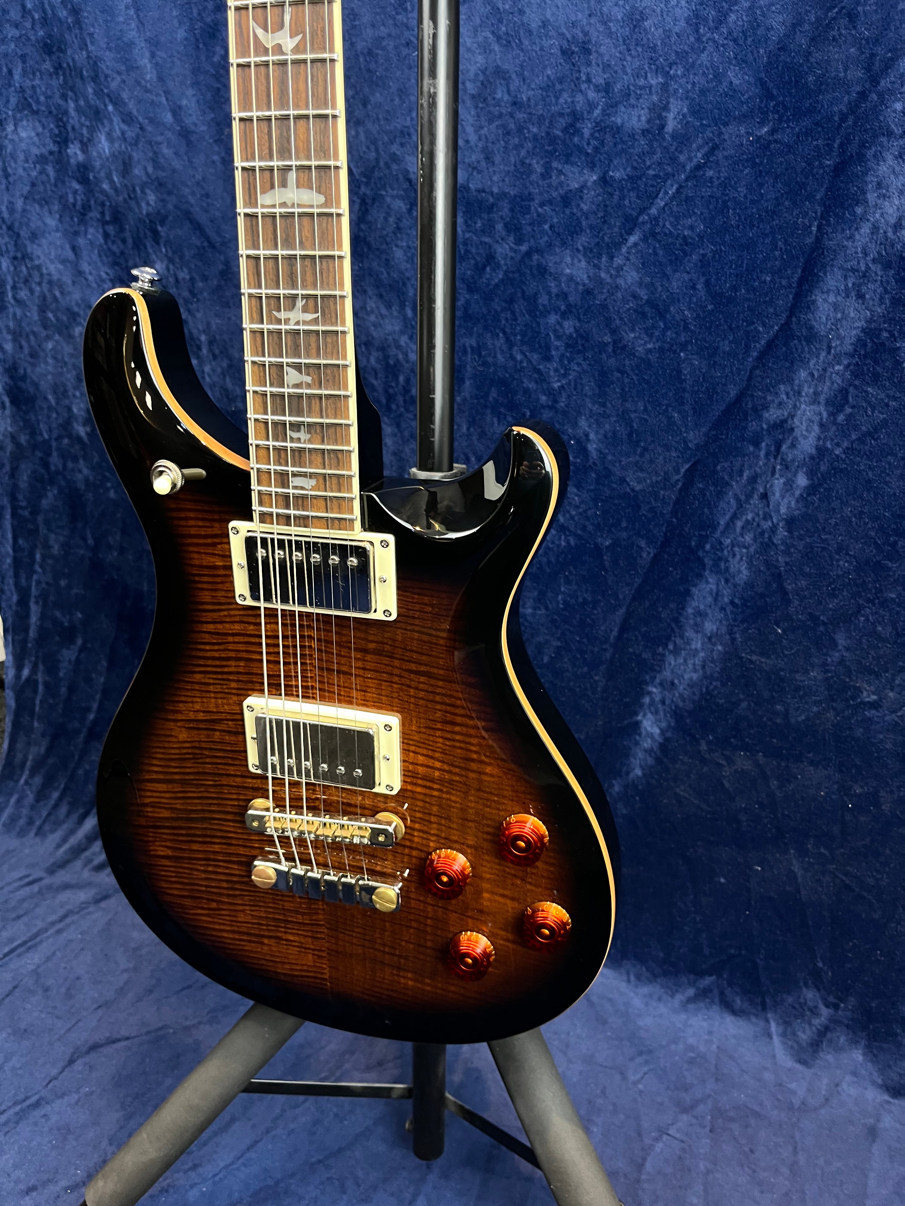 Sunburst electric guitar with two pickups, four orange control knobs, and bird-shaped inlays on the fretboard, shown on a stand against a blue backdrop.