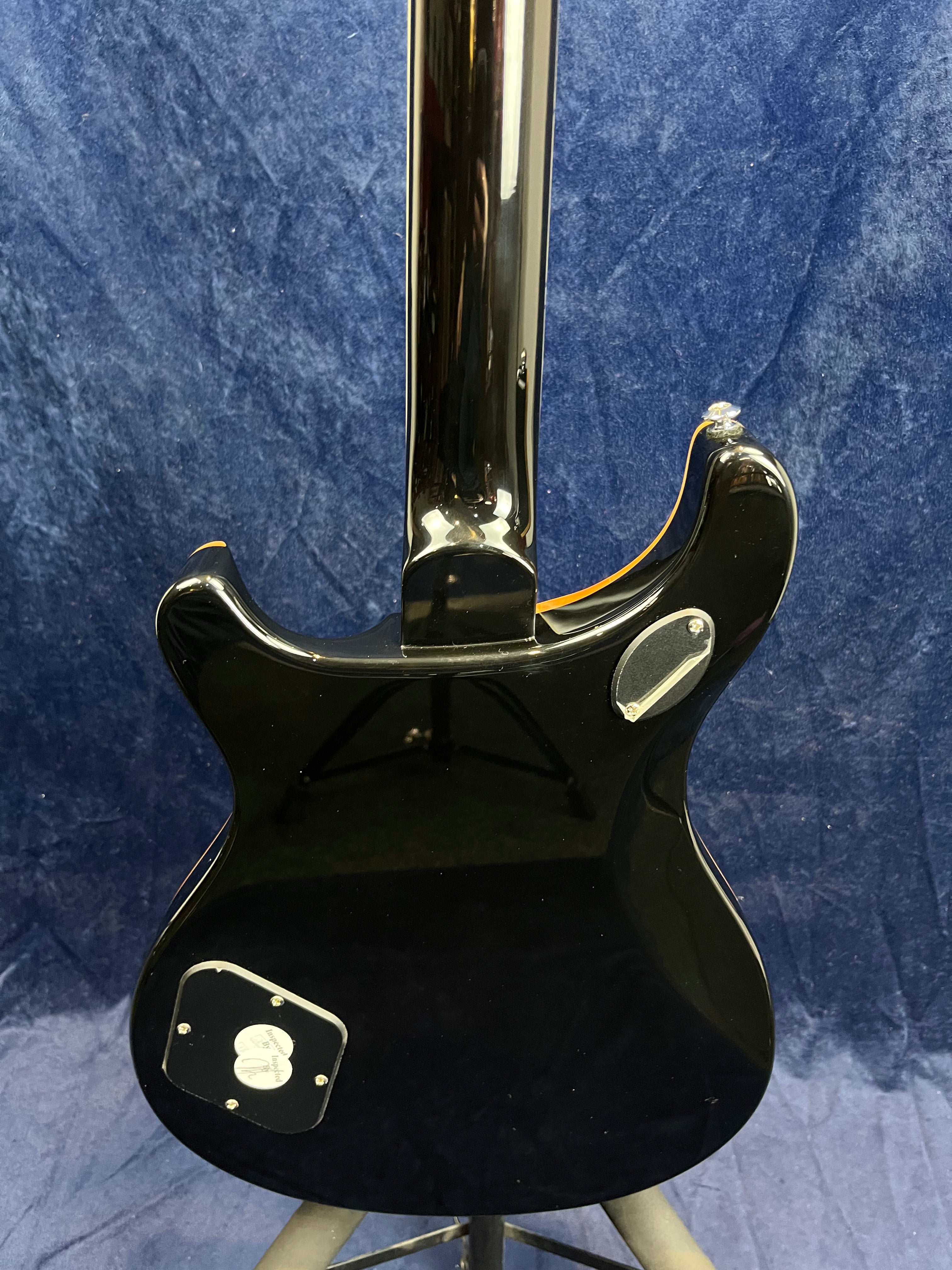 Glossy black electric guitar shown from the back with chrome hardware and a control cavity cover near the lower edge, against a blue fabric backdrop.