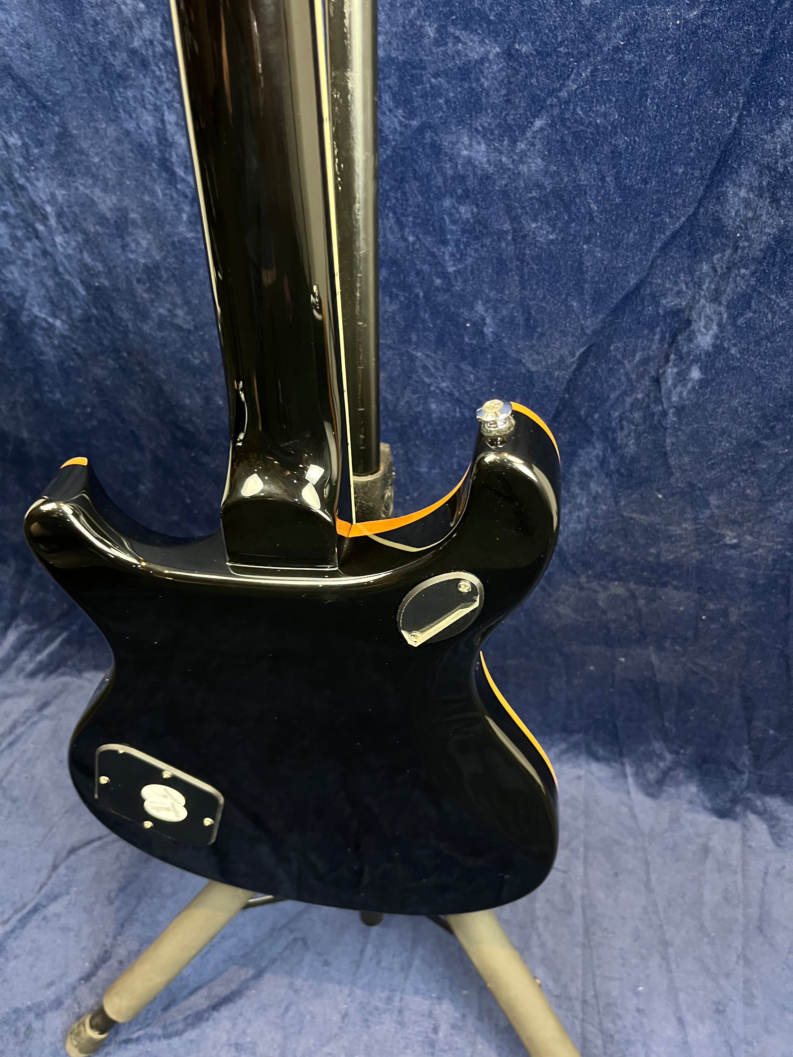 Black electric guitar shown from the back, with a glossy finish, orange edge trim, and mounted on a tripod stand against a dark blue background.