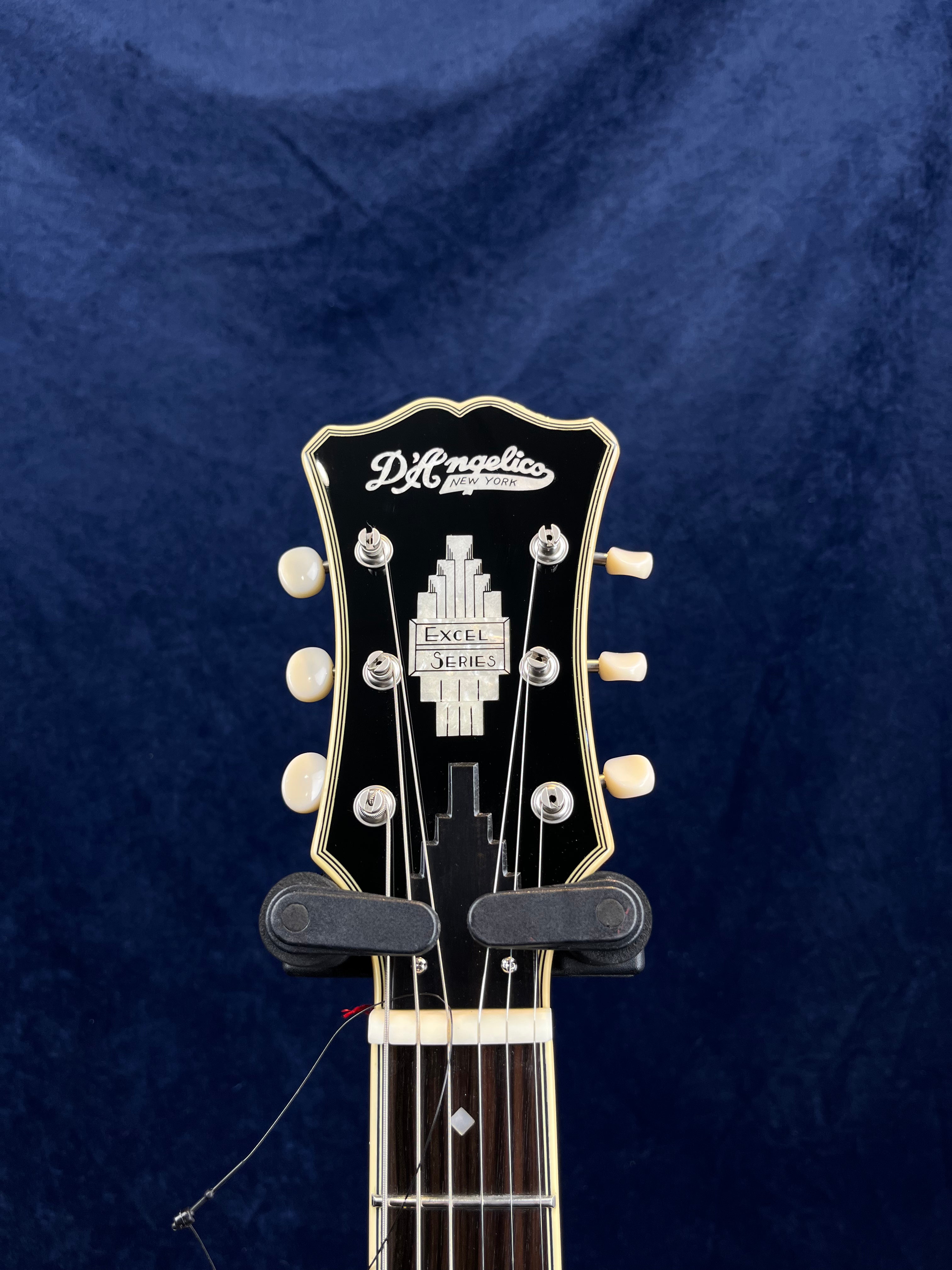 Headstock of a D’Angelico Excel Series electric guitar, black with cream trim, six cream tuning pegs, and art deco inlay design.
