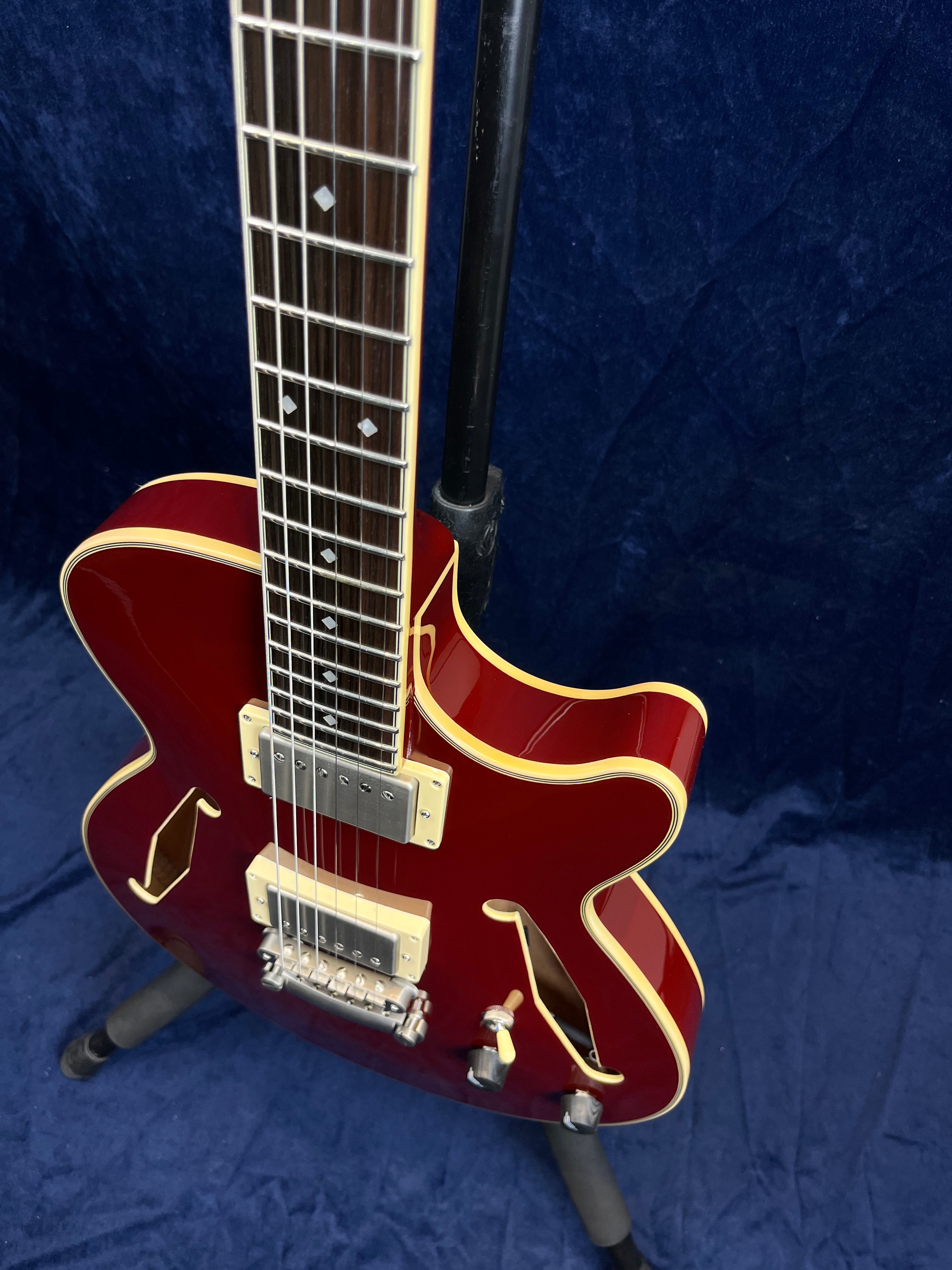 Red semi-hollow electric guitar with cream binding, two humbucker pickups, f-holes, and chrome hardware, shown standing on a tripod stand.
