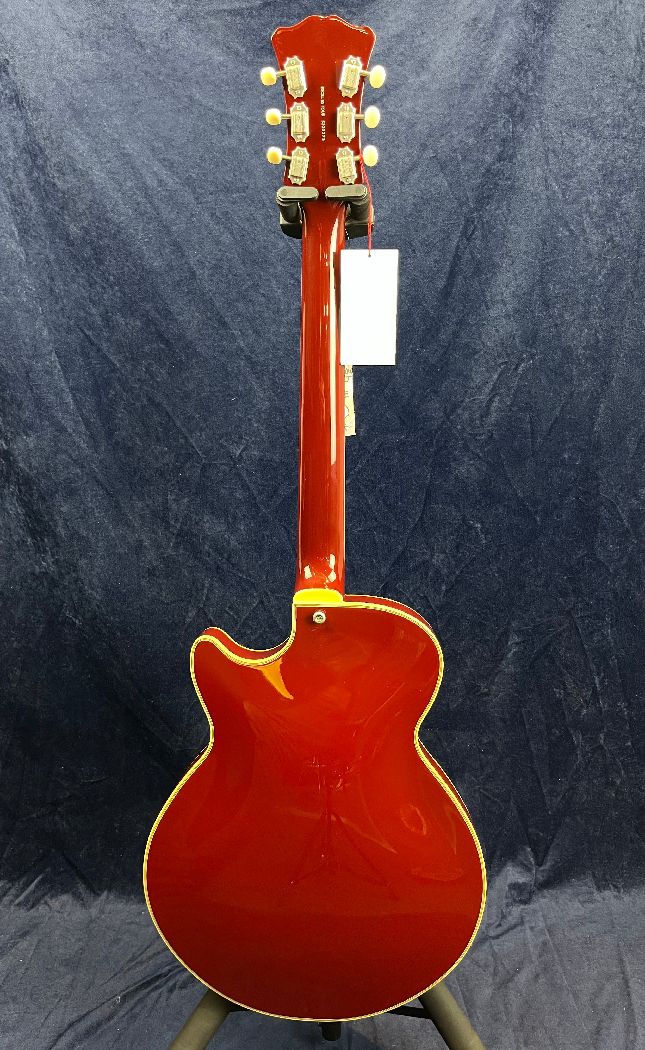 Glossy red electric guitar shown from the back, yellow binding around the edges, six cream tuning pegs, and a white tag hanging from the headstock.