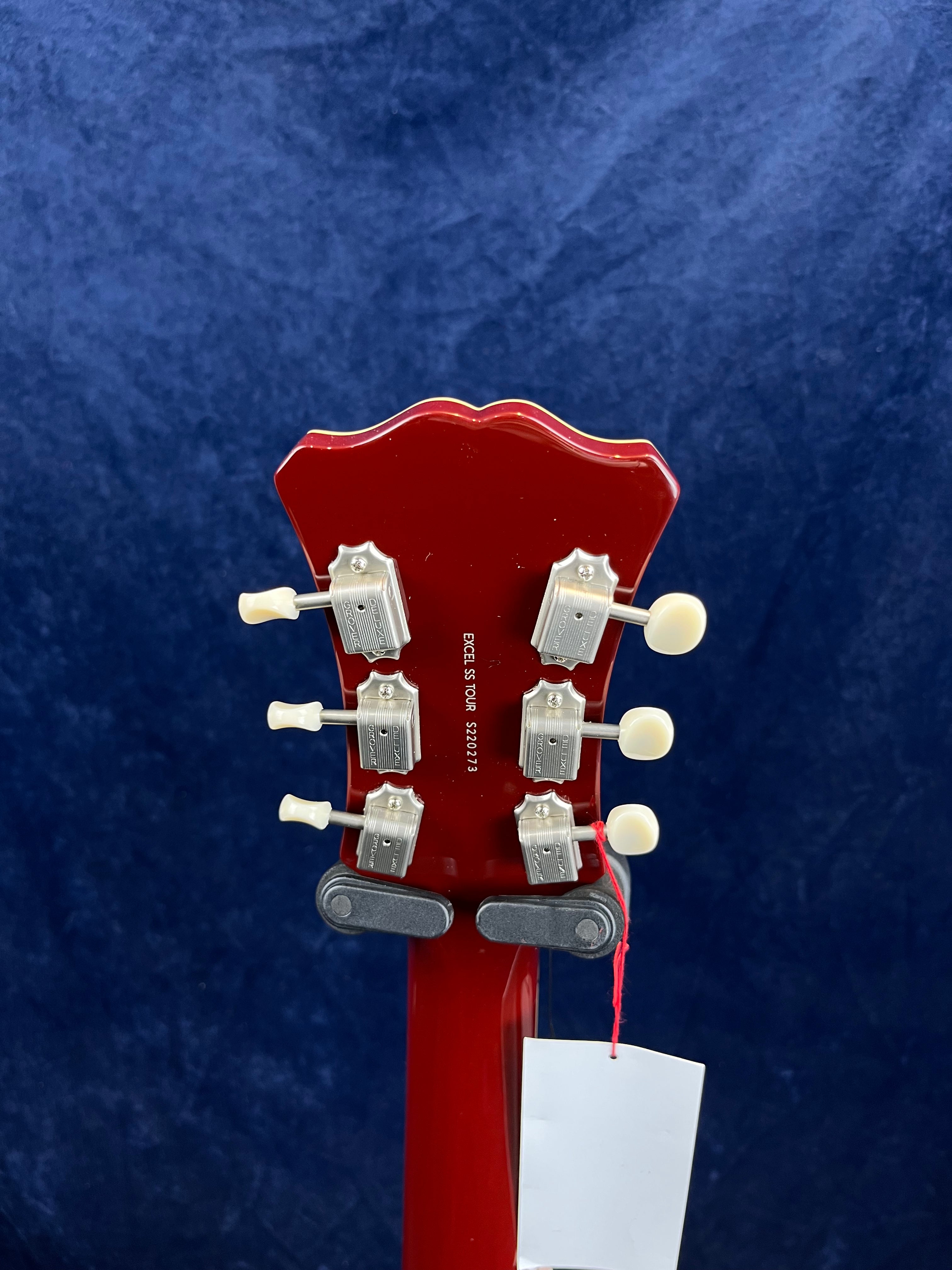 Back of a red electric guitar headstock with six silver tuning pegs and cream knobs against a dark blue background; a white tag hangs below.