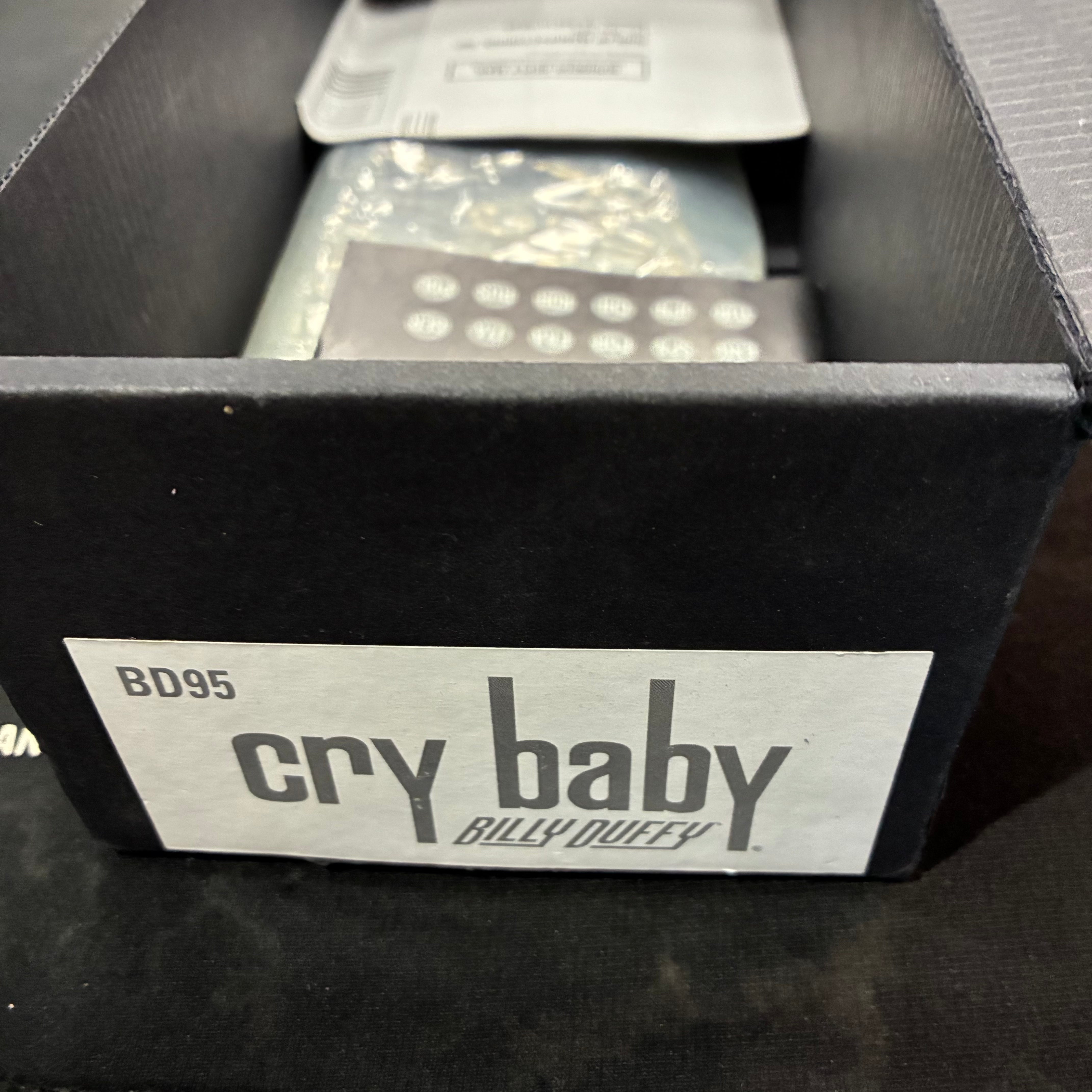 A black FX pedal box labeled "BD95 cry baby Billy Duffy," with paperwork and the pedal visible inside the open box.