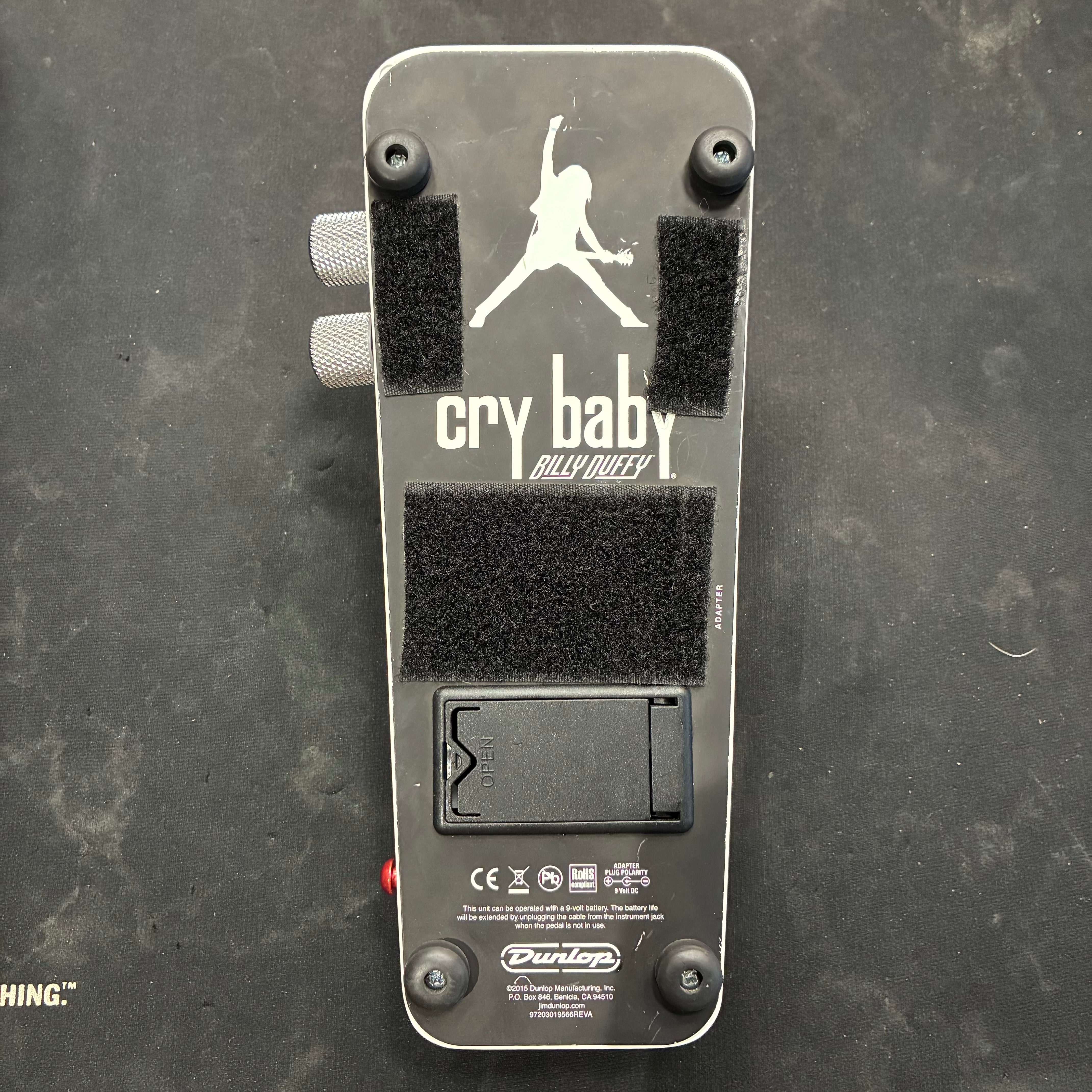The bottom of a Dunlop Cry Baby Billy Duffy FX pedal with battery compartment, velcro strips, and a silhouette graphic of a person playing guitar.