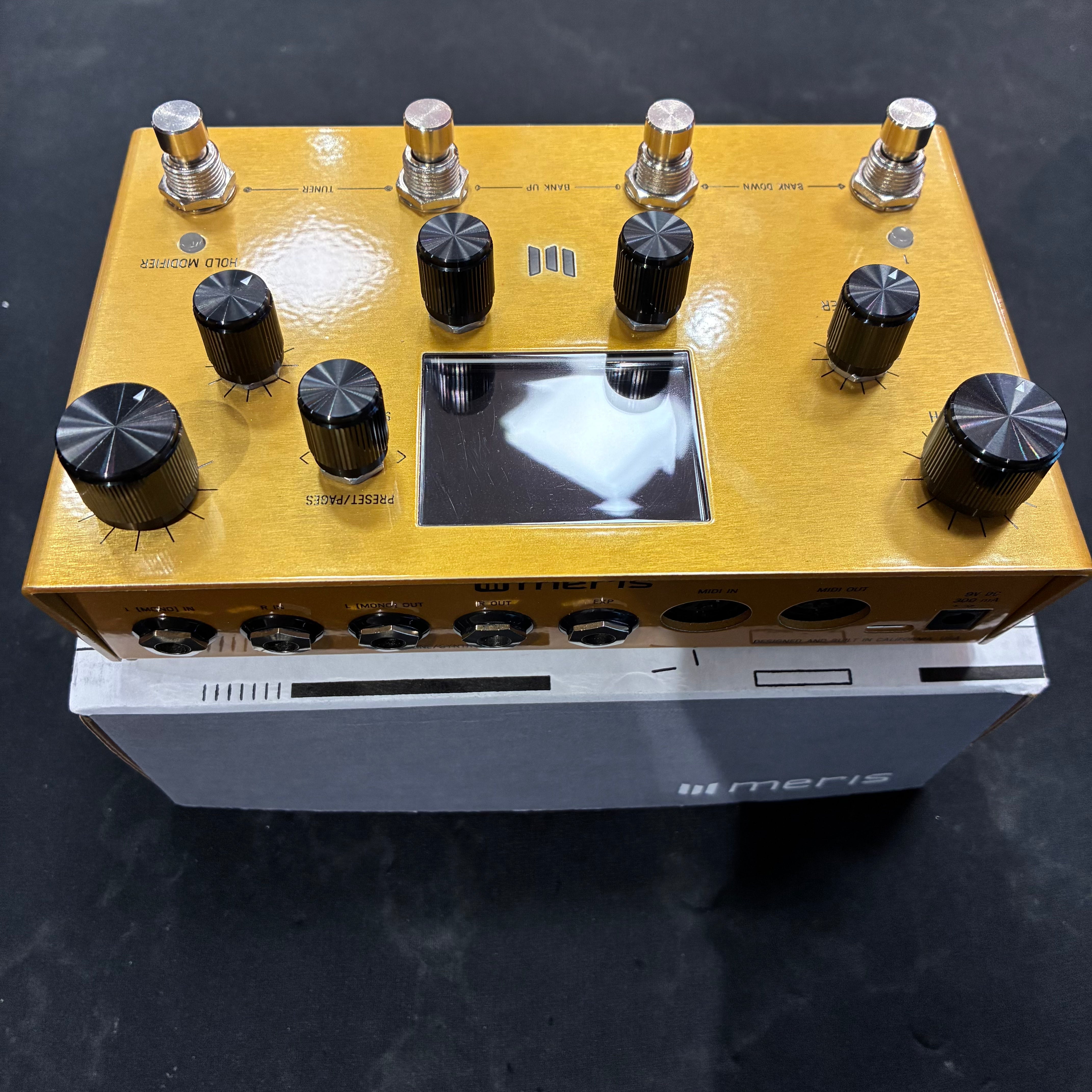 Gold Meris FX pedal with seven black knobs, three footswitches, and a central display screen, sitting on a grey Meris box.