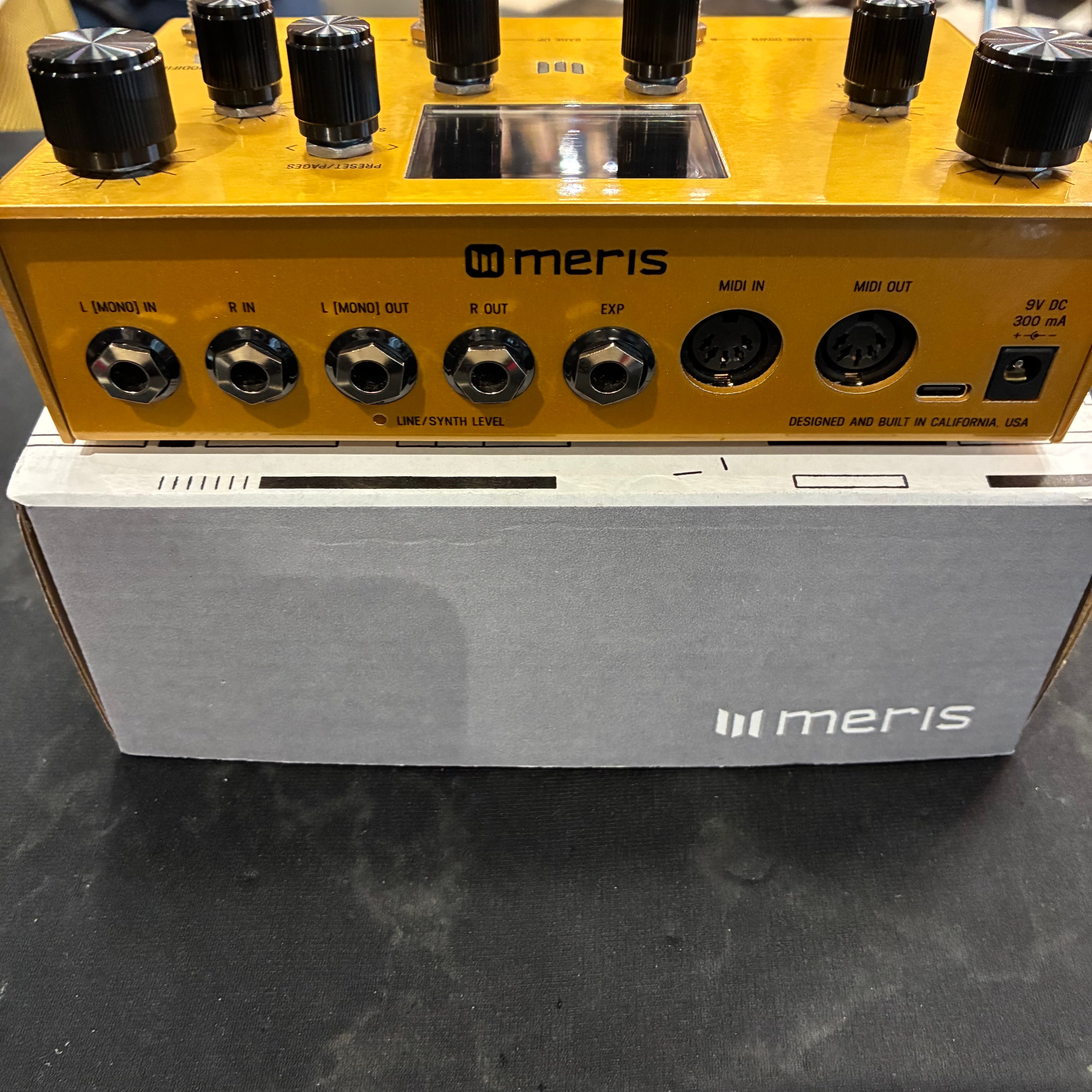 Golden Meris FX pedal showing multiple input, output, expression, MIDI, and power ports, resting on a gray Meris-branded box.