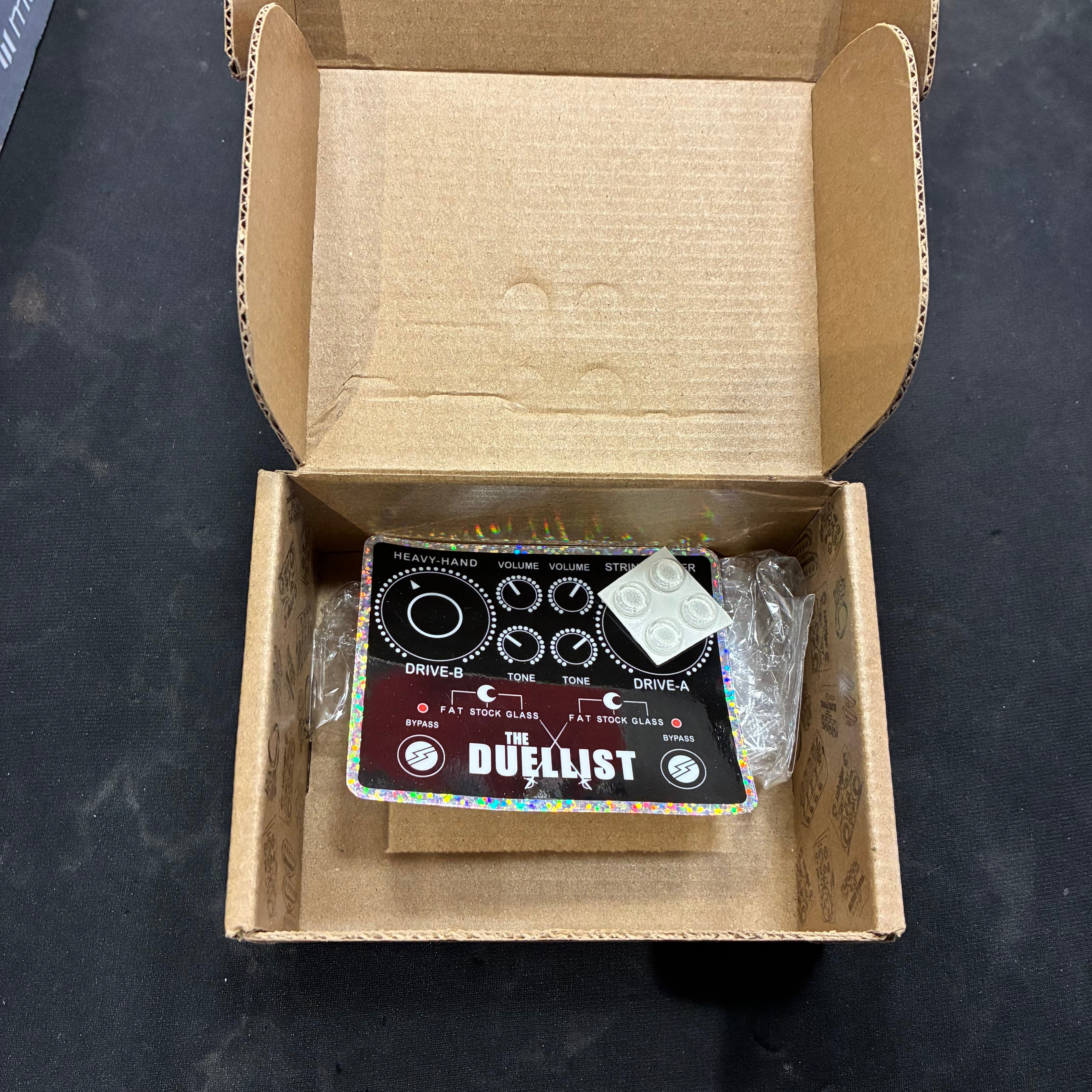 FX pedal called The Duellist inside an open cardboard box, showing black faceplate with multiple knobs and white label dots nearby.