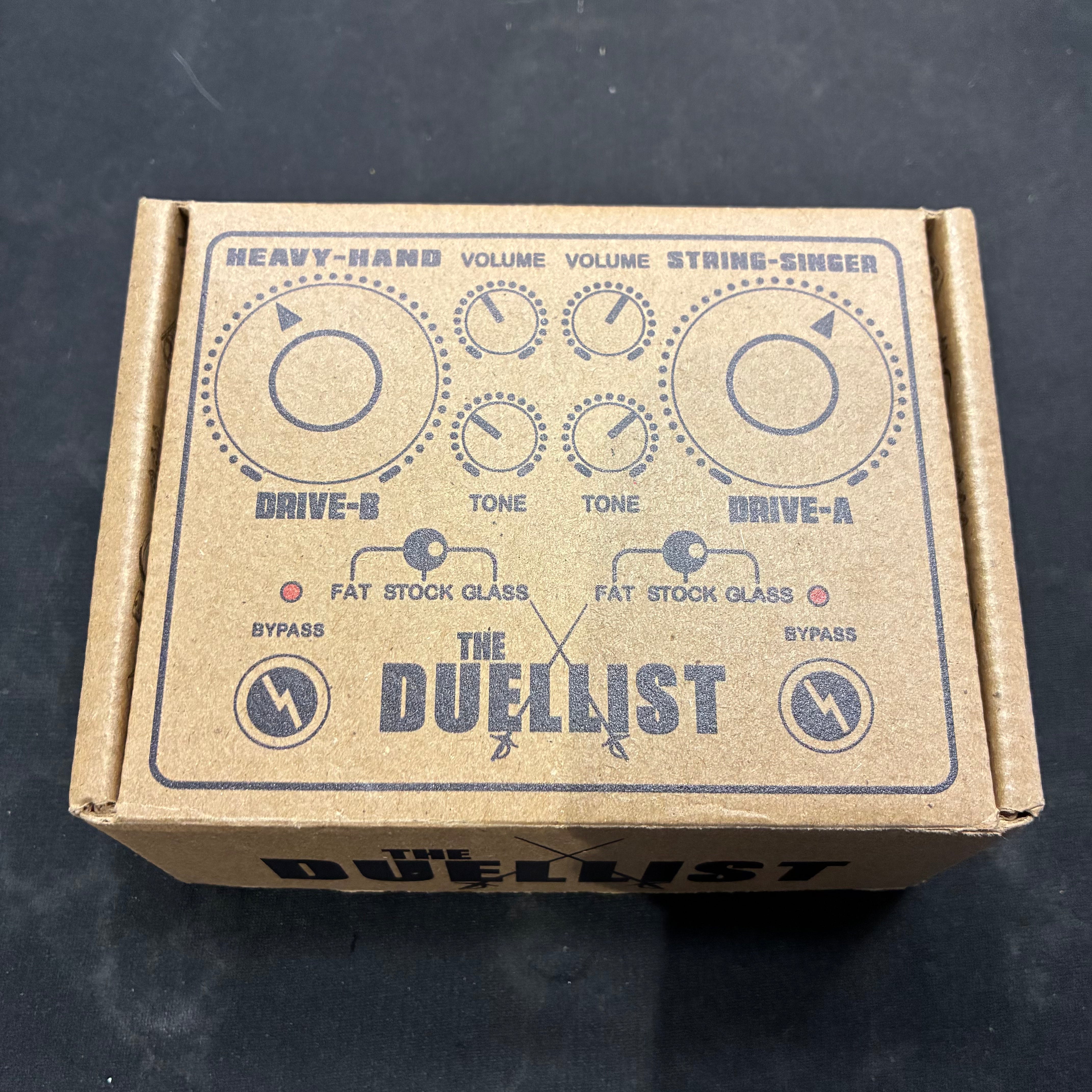 Cardboard box featuring a diagram of The Duellist FX pedal, showing controls for Drive-A, Drive-B, tone knobs, volume knobs, and mode switches.