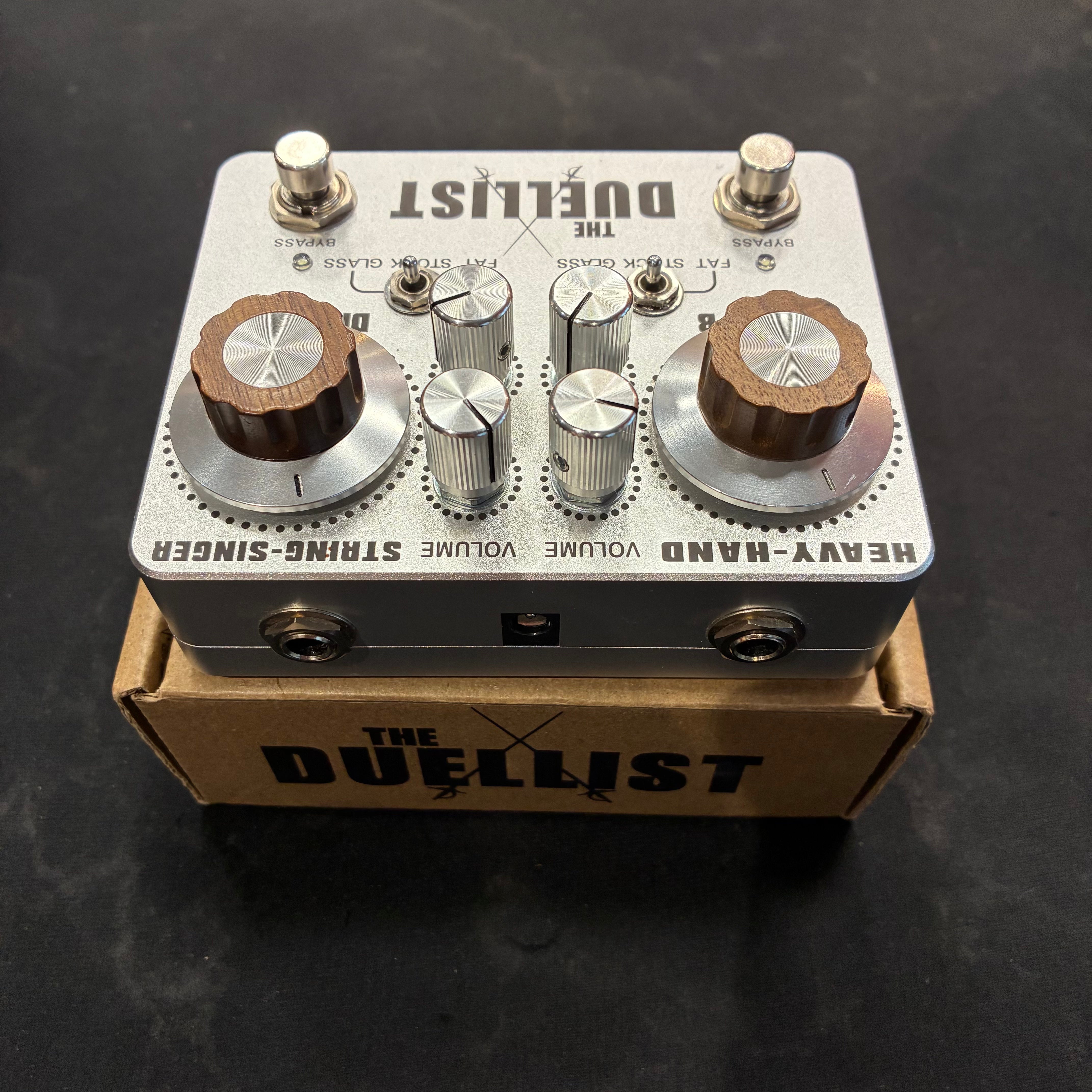 Silver Duelist FX pedal with two footswitches, seven control knobs, and toggle switches, sitting on a branded cardboard box.