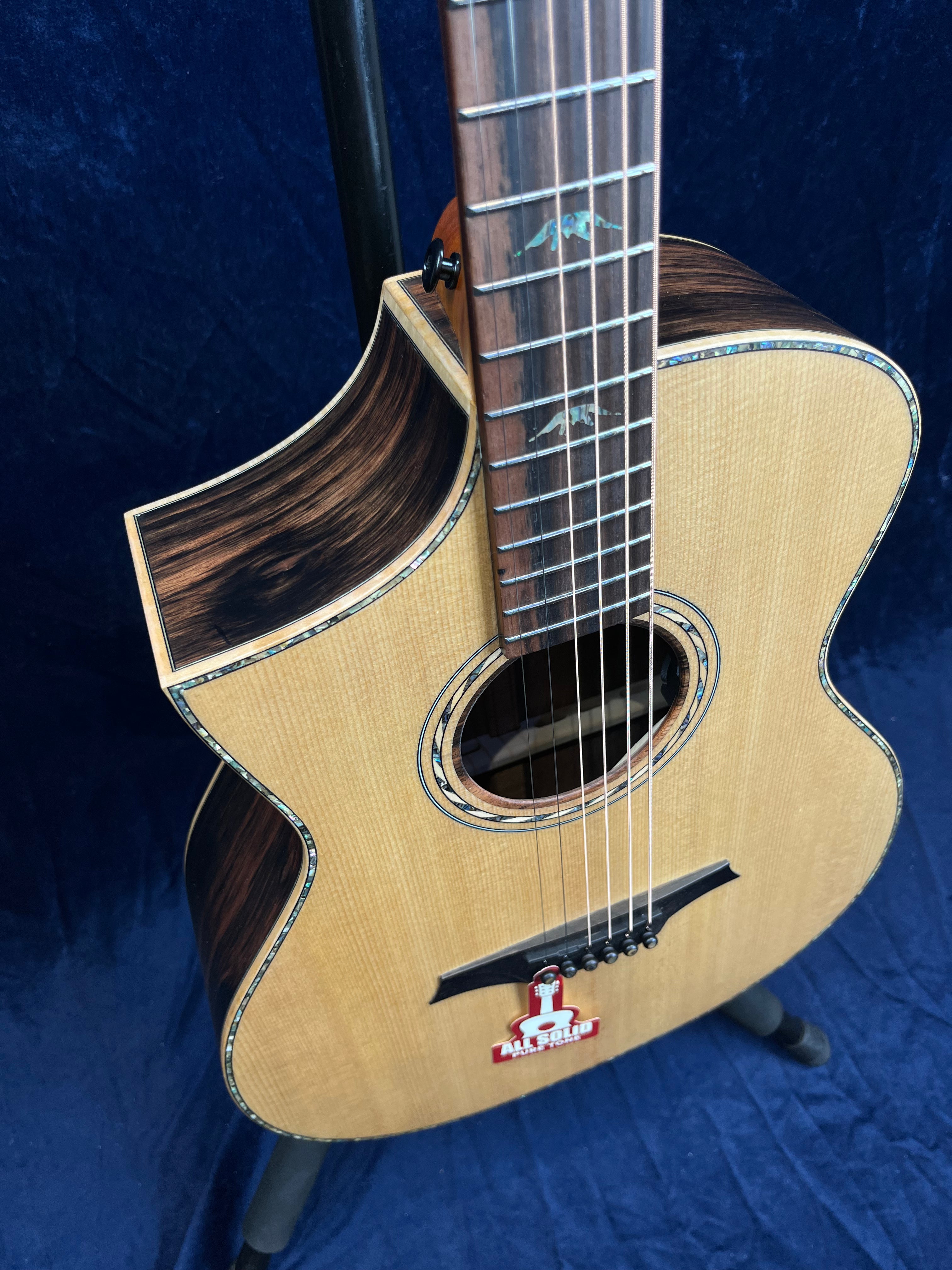 Acoustic guitar with a natural wood top, dark wood accents, and abalone inlays around the soundhole and fretboard, displayed upright on a stand.