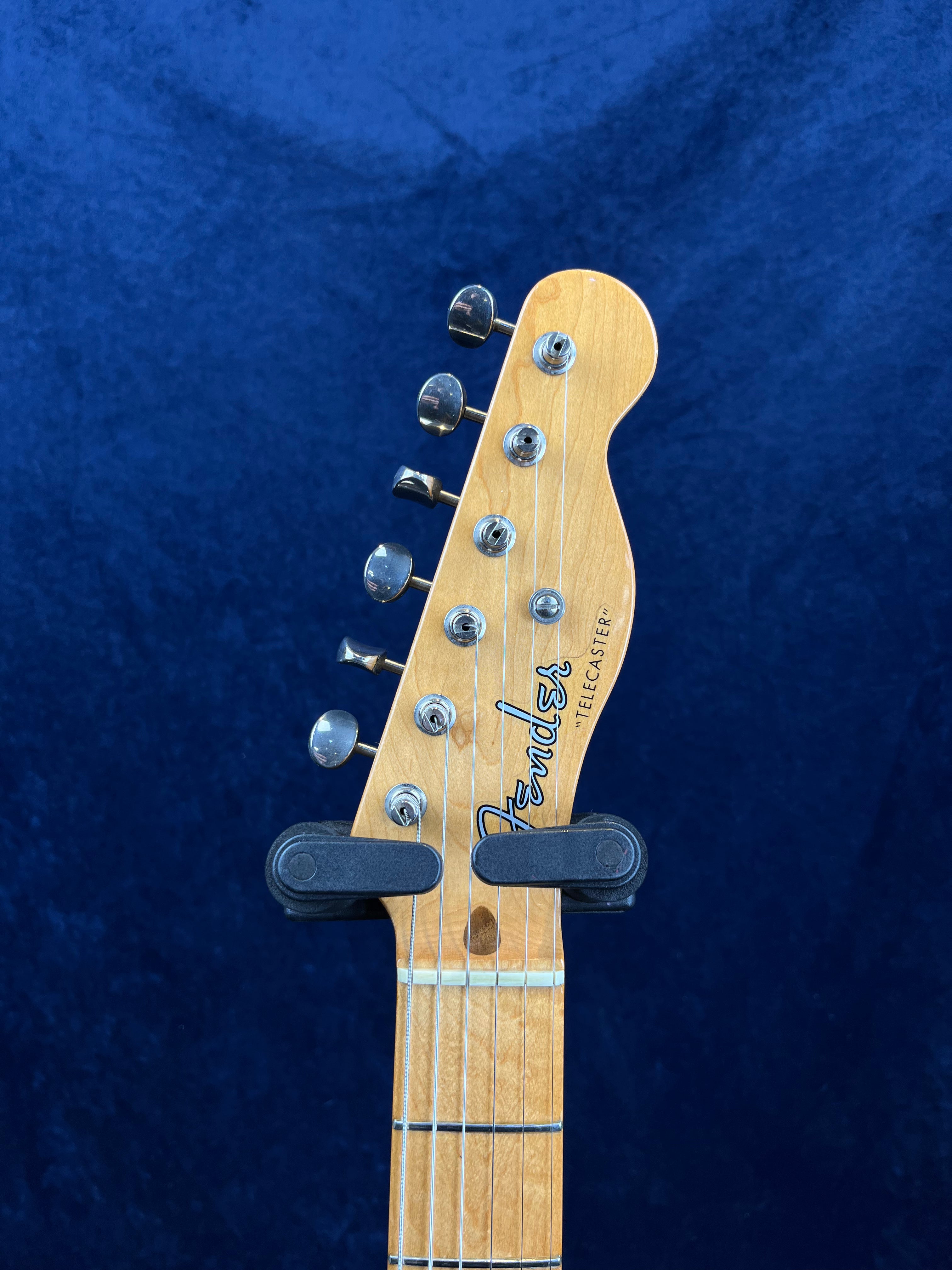 Light wood Fender Telecaster electric guitar headstock with six tuning pegs, logo, and capo clipped just below the headstock against a dark blue background.