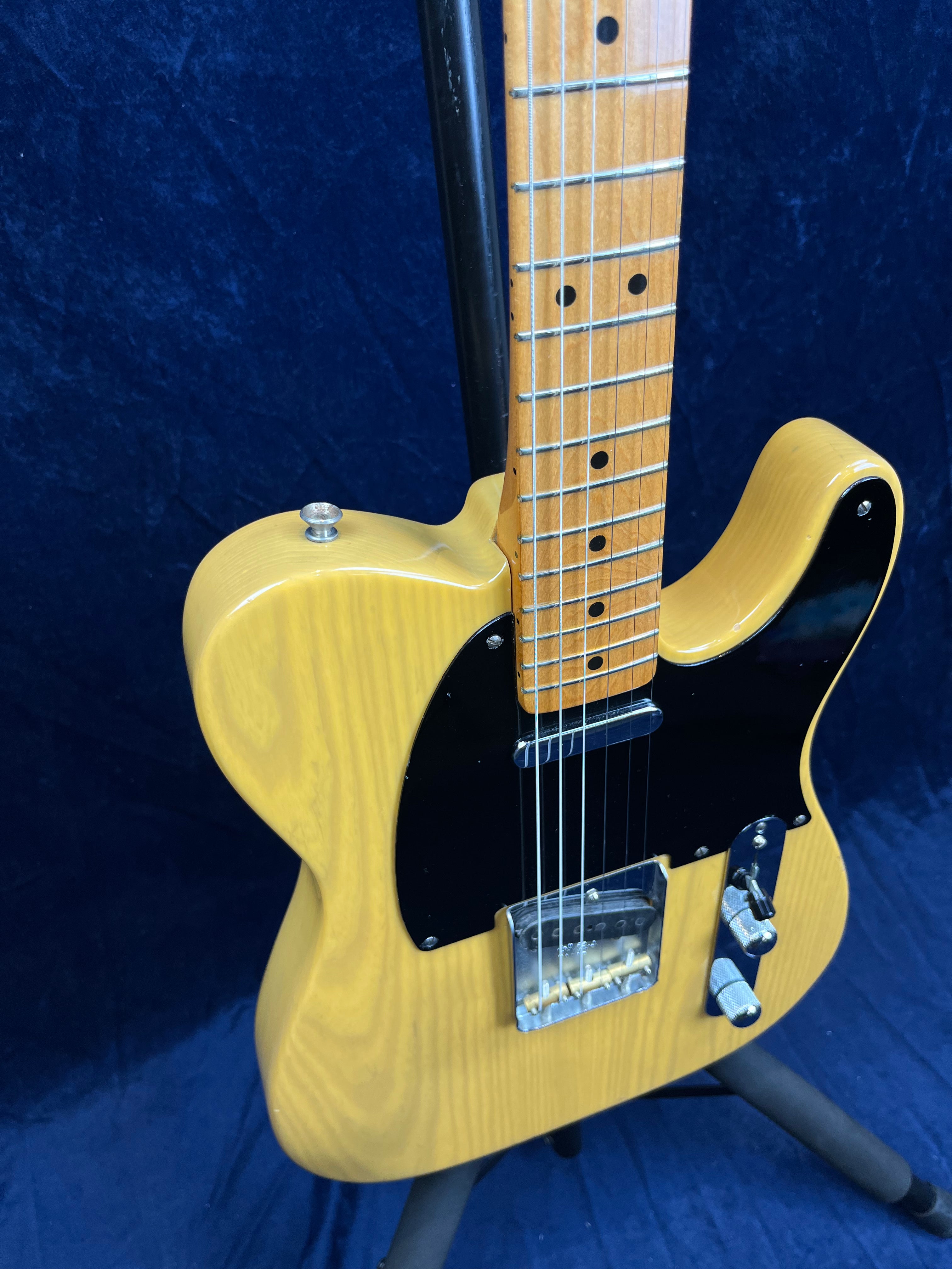 Light wood electric guitar with black pickguard, two metal control knobs, and six strings, shown on a stand against a dark blue background.