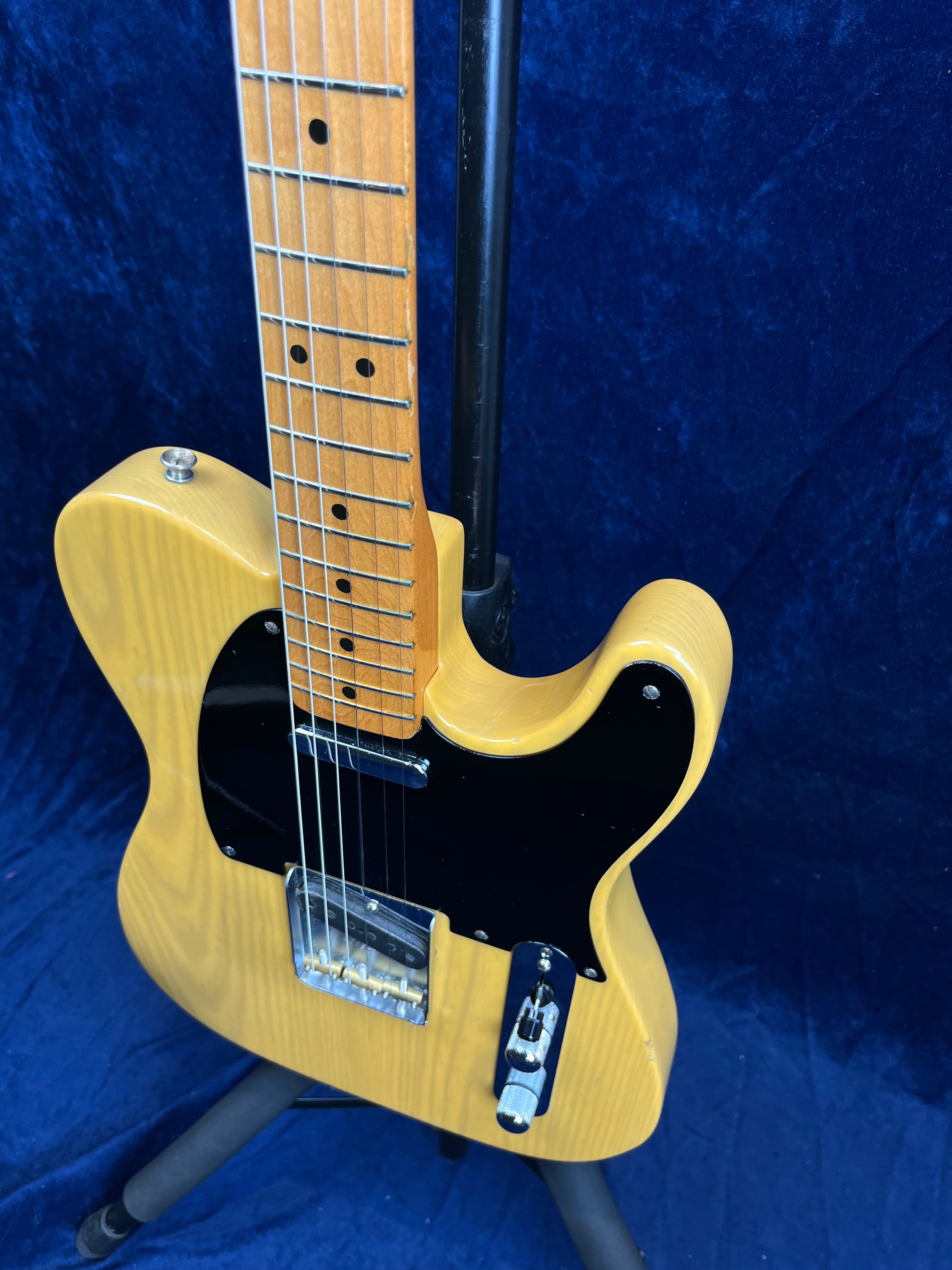 Blonde-finished electric guitar with black pickguard, maple neck, and chrome hardware, shown upright on a stand against a dark blue background.