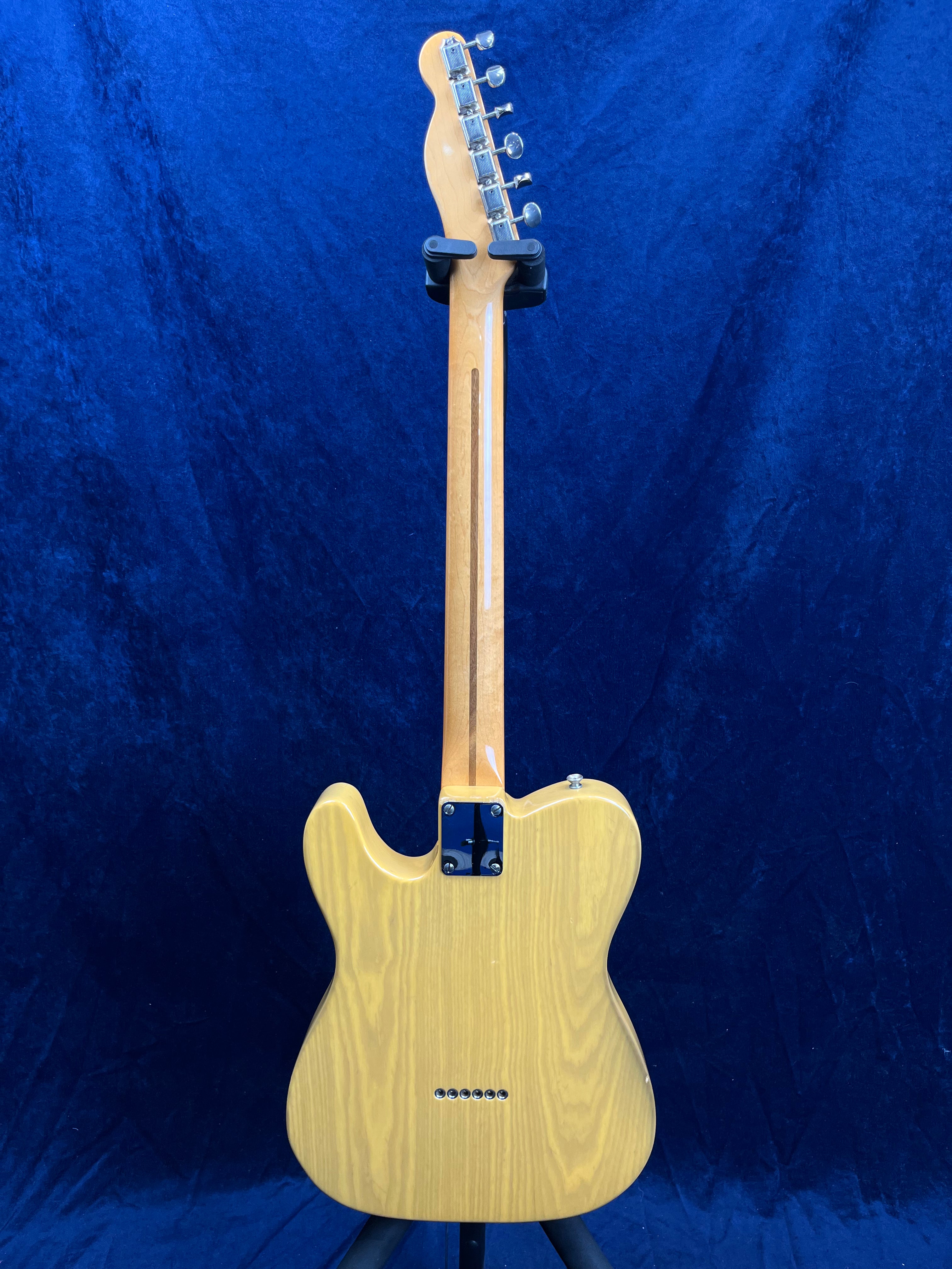 Light wood electric guitar shown from the back, displaying its neck, tuning pegs, and smooth grain body against a blue background.