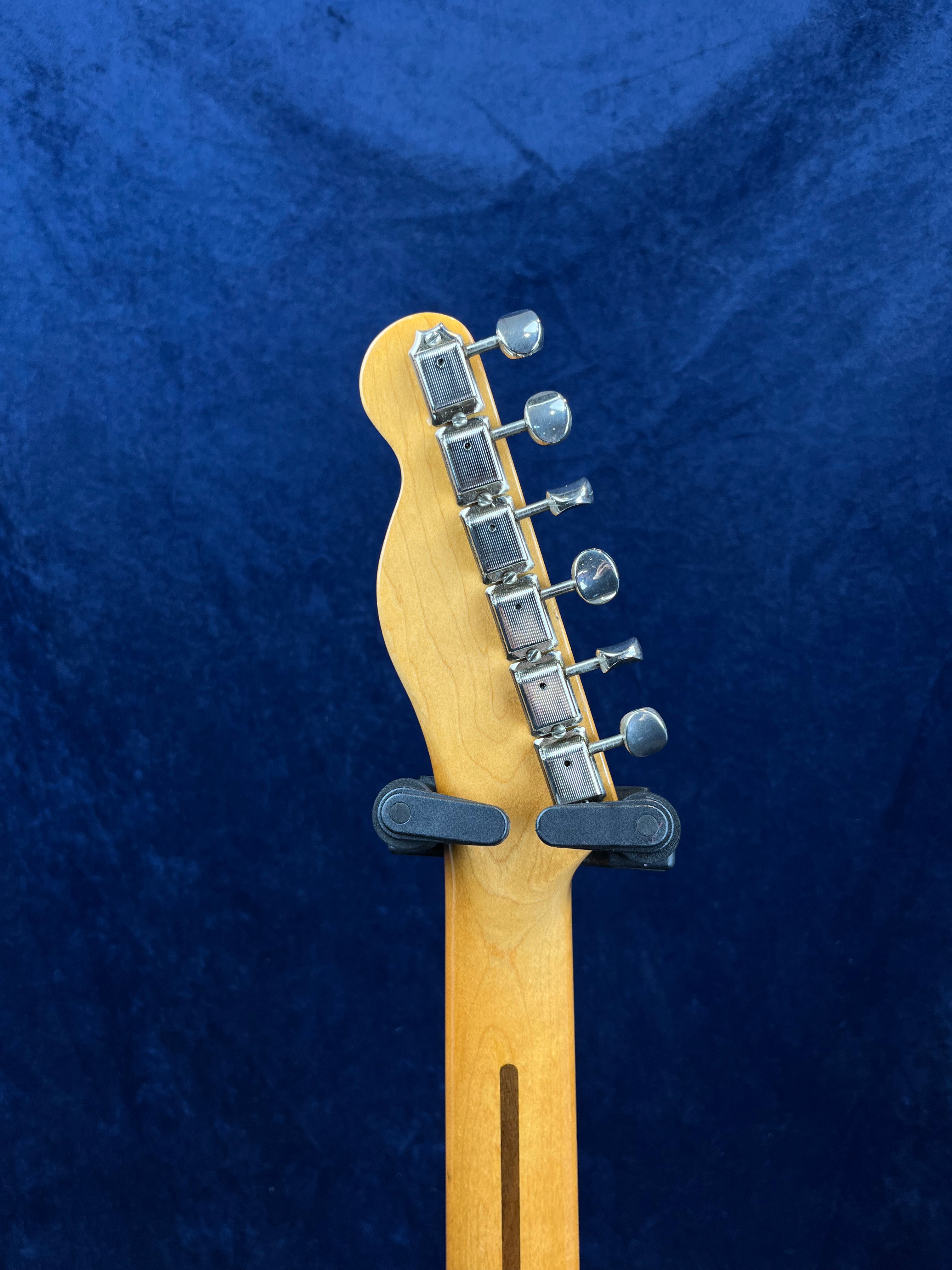 The back of an electric guitar headstock with six metal tuning pegs, light wood finish, and a dark blue background.