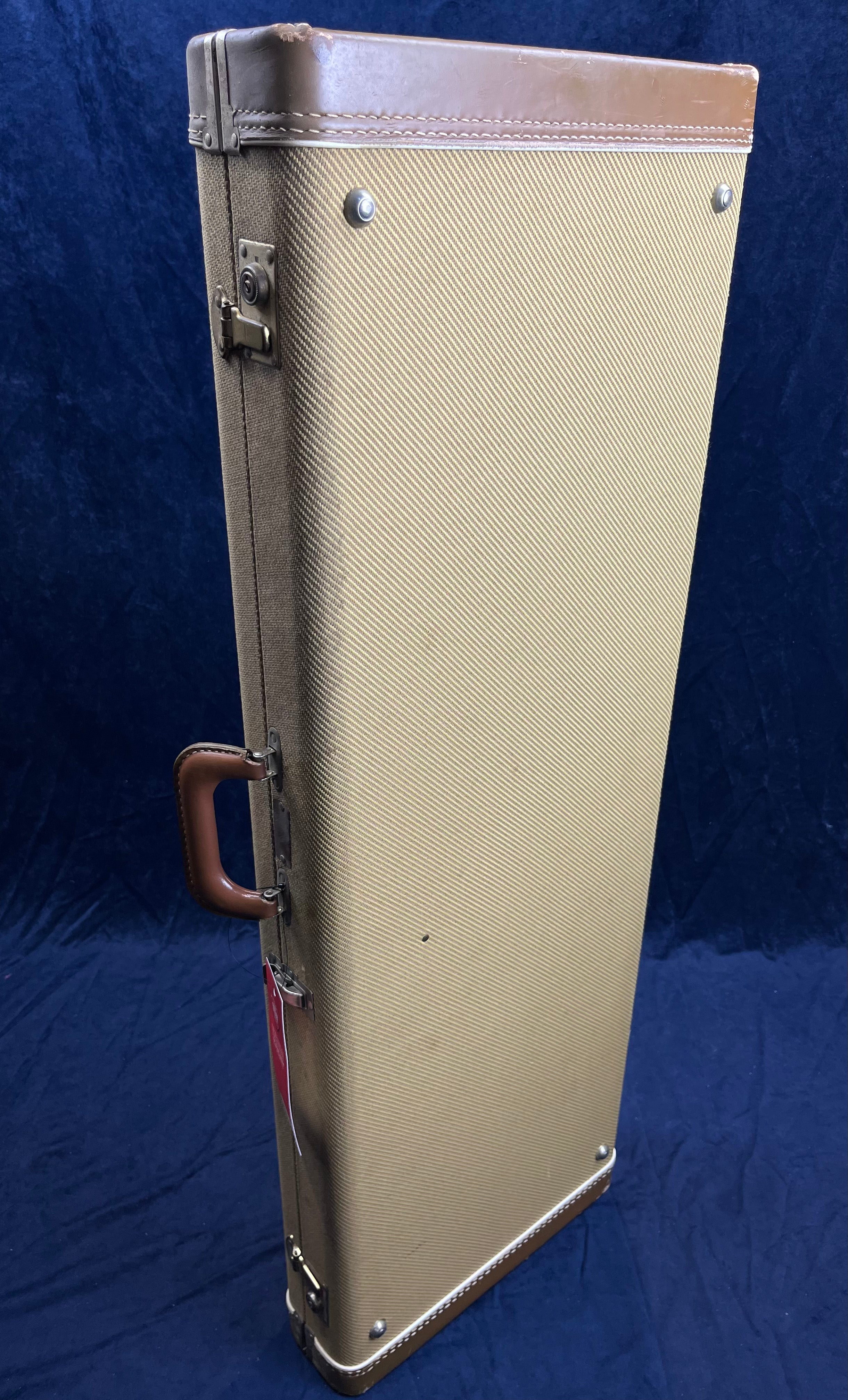 Rectangular tan and brown electric guitar case stands upright, with a leather handle, metal latches, and protective metal corners.
