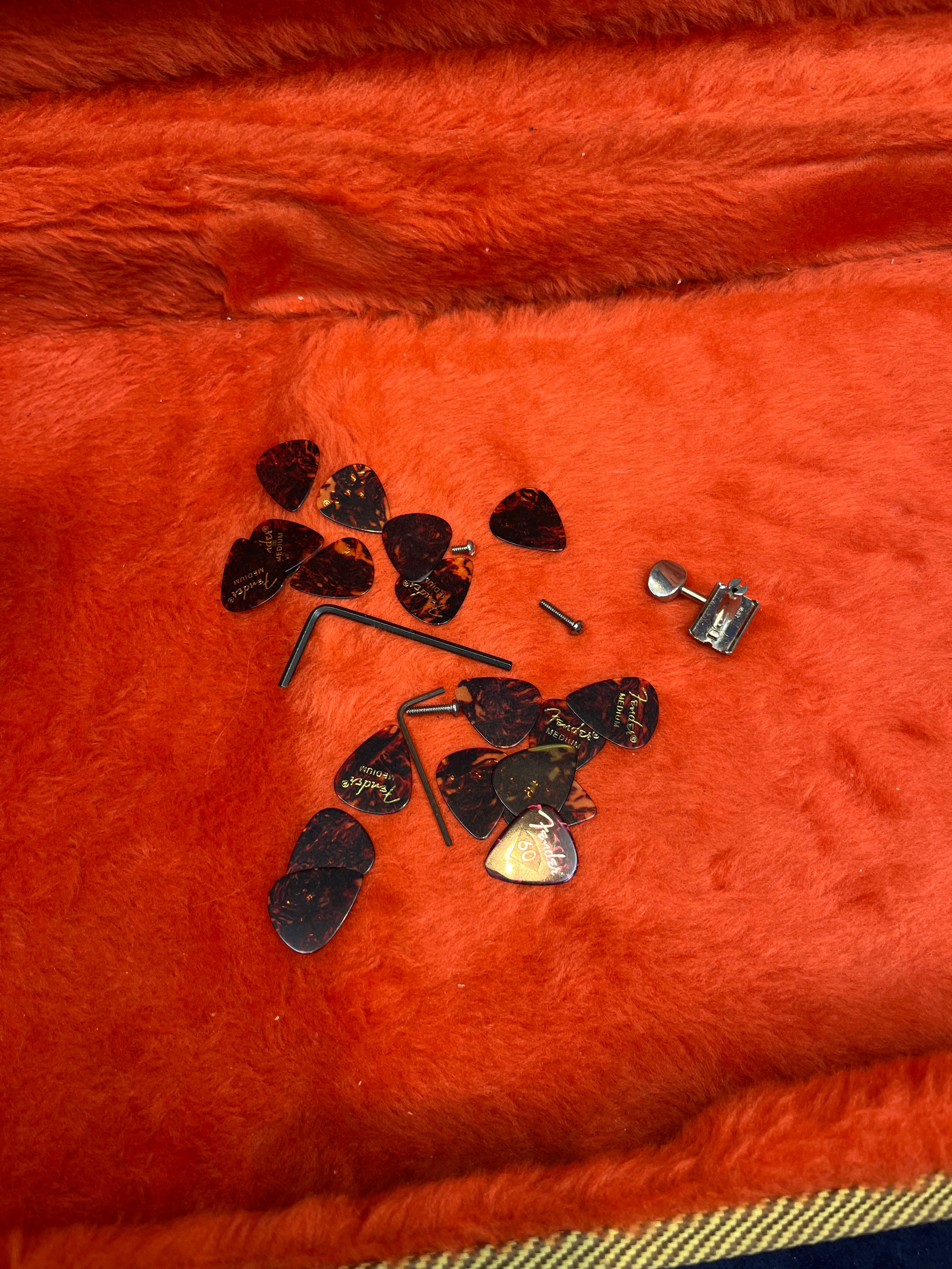 Several guitar picks, a tuning peg, some small screws, and two hex wrenches lie on orange fuzzy fabric inside an electric guitar case.
