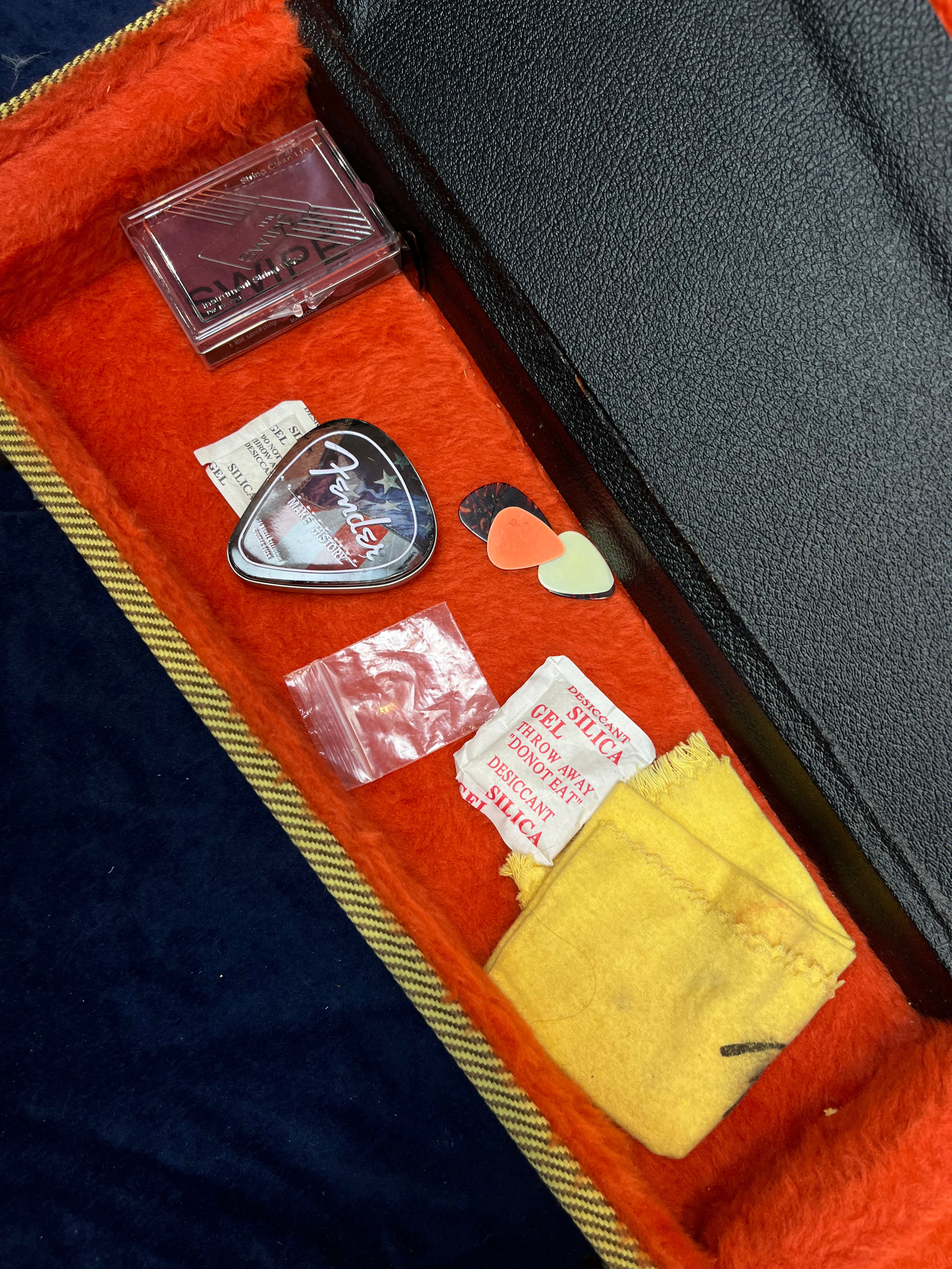 Electric guitar case compartment with guitar picks, a Fender-branded tin, silica gel packets, a yellow cloth, and a plastic container.