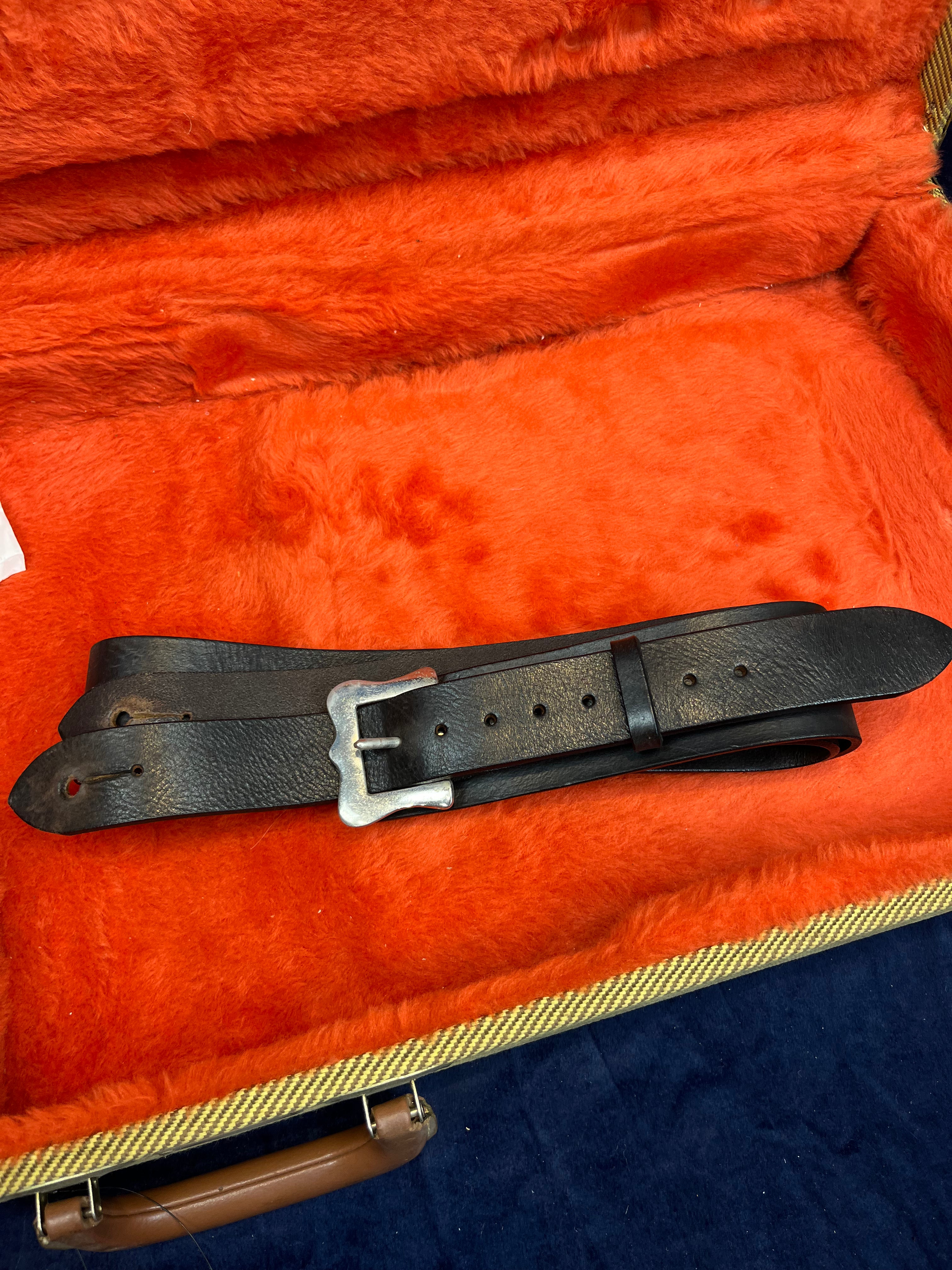 A black leather guitar strap with a metal buckle rests inside an orange plush-lined electric guitar case.