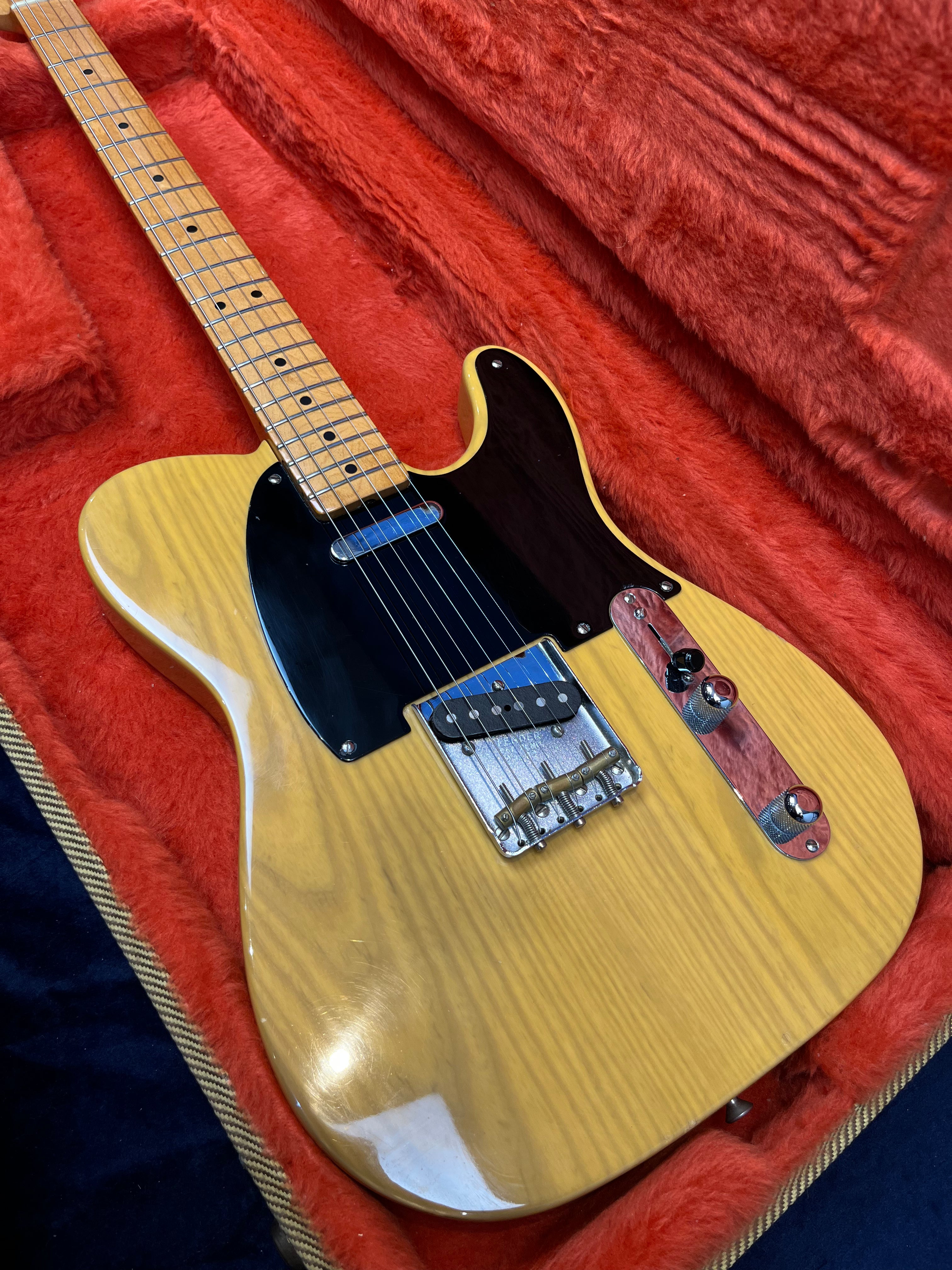 Blonde wood electric guitar with black pickguard, chrome bridge, and control plate, resting in a plush red-lined hard case.
