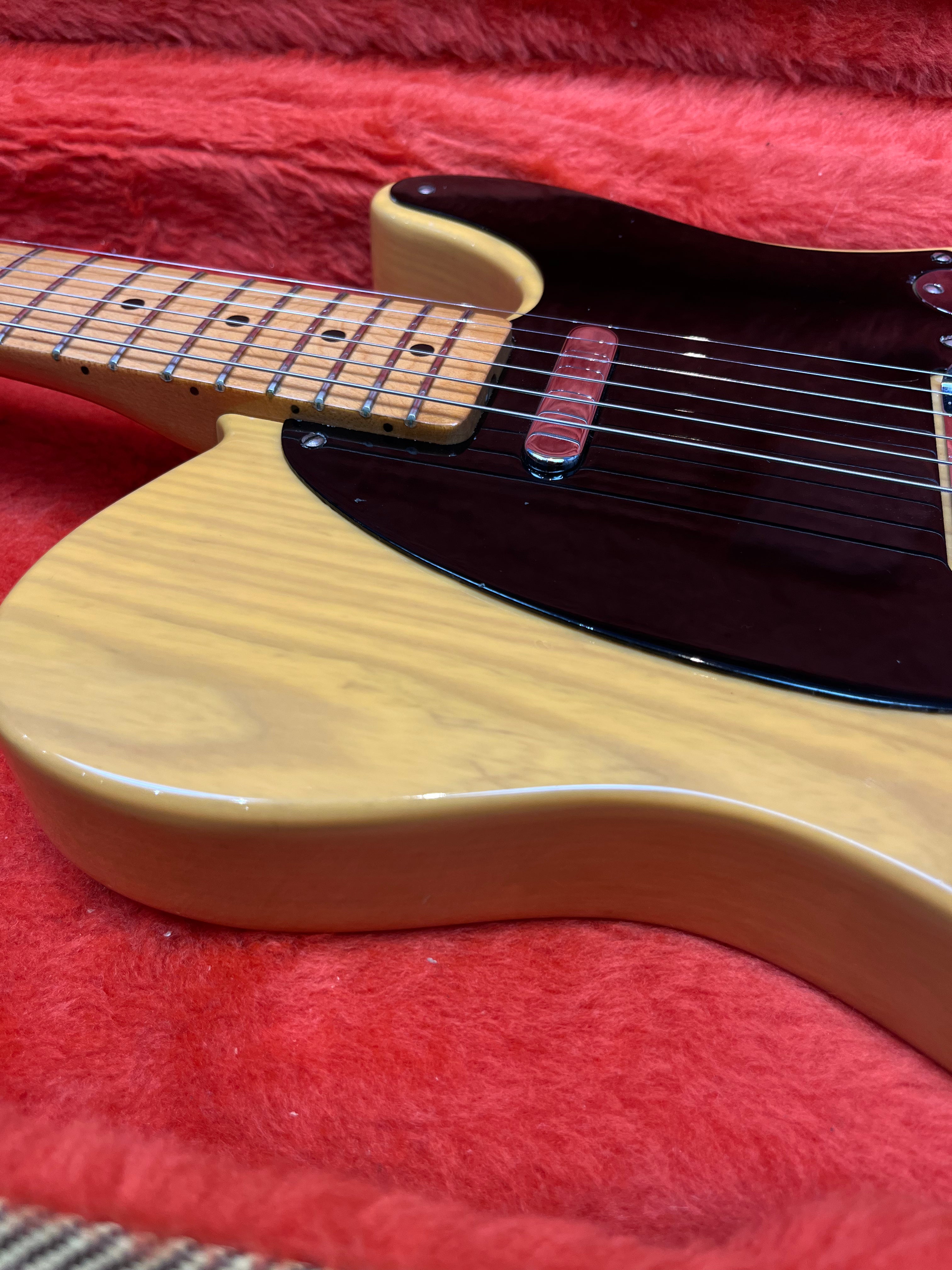Light wood electric guitar with a maple neck, black pickguard, and chrome pickup resting in a red plush-lined case.