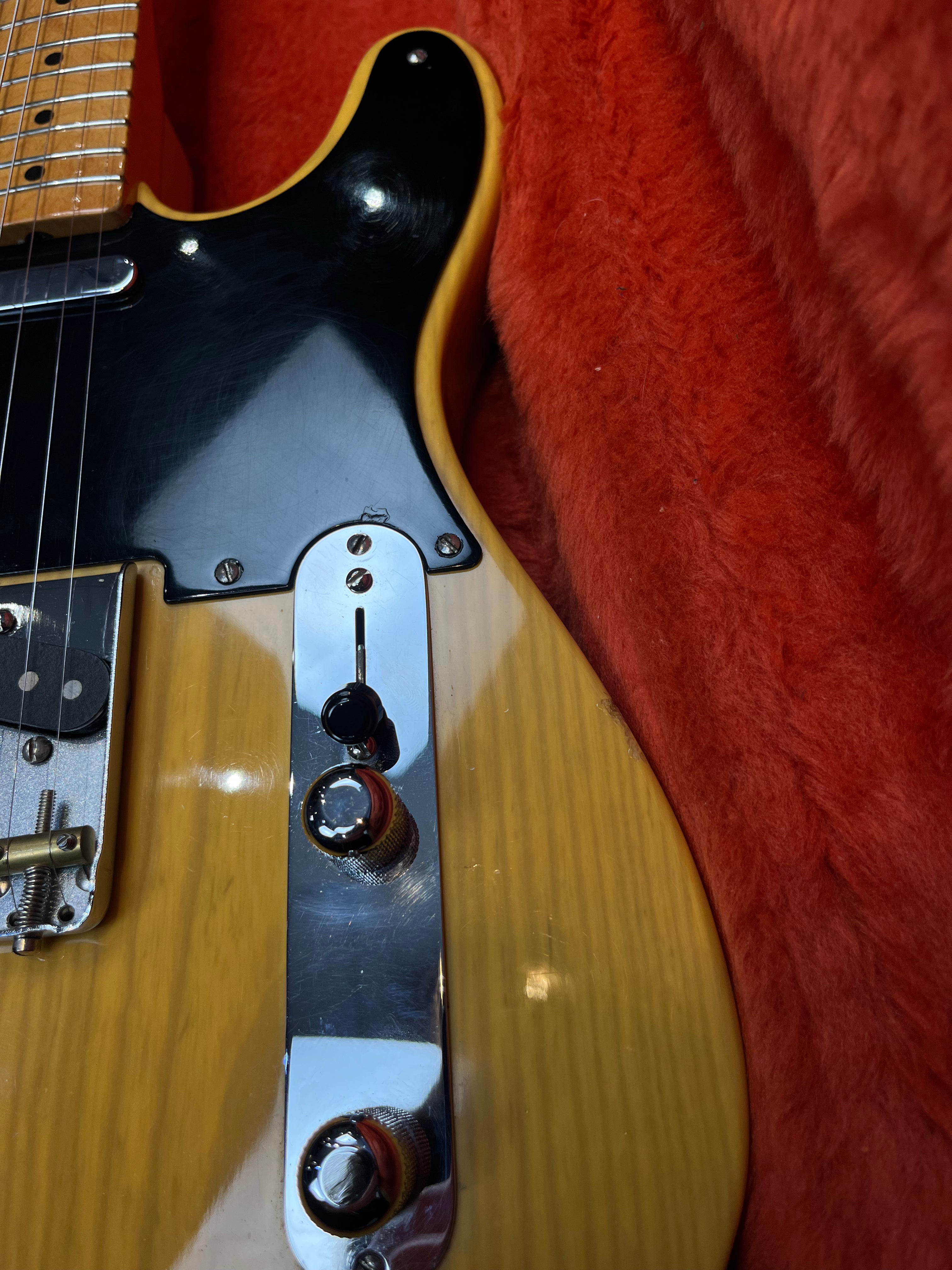 Close-up of a yellow electric guitar body with a black pickguard, two control knobs, and a chrome switch panel against a red plush background.
