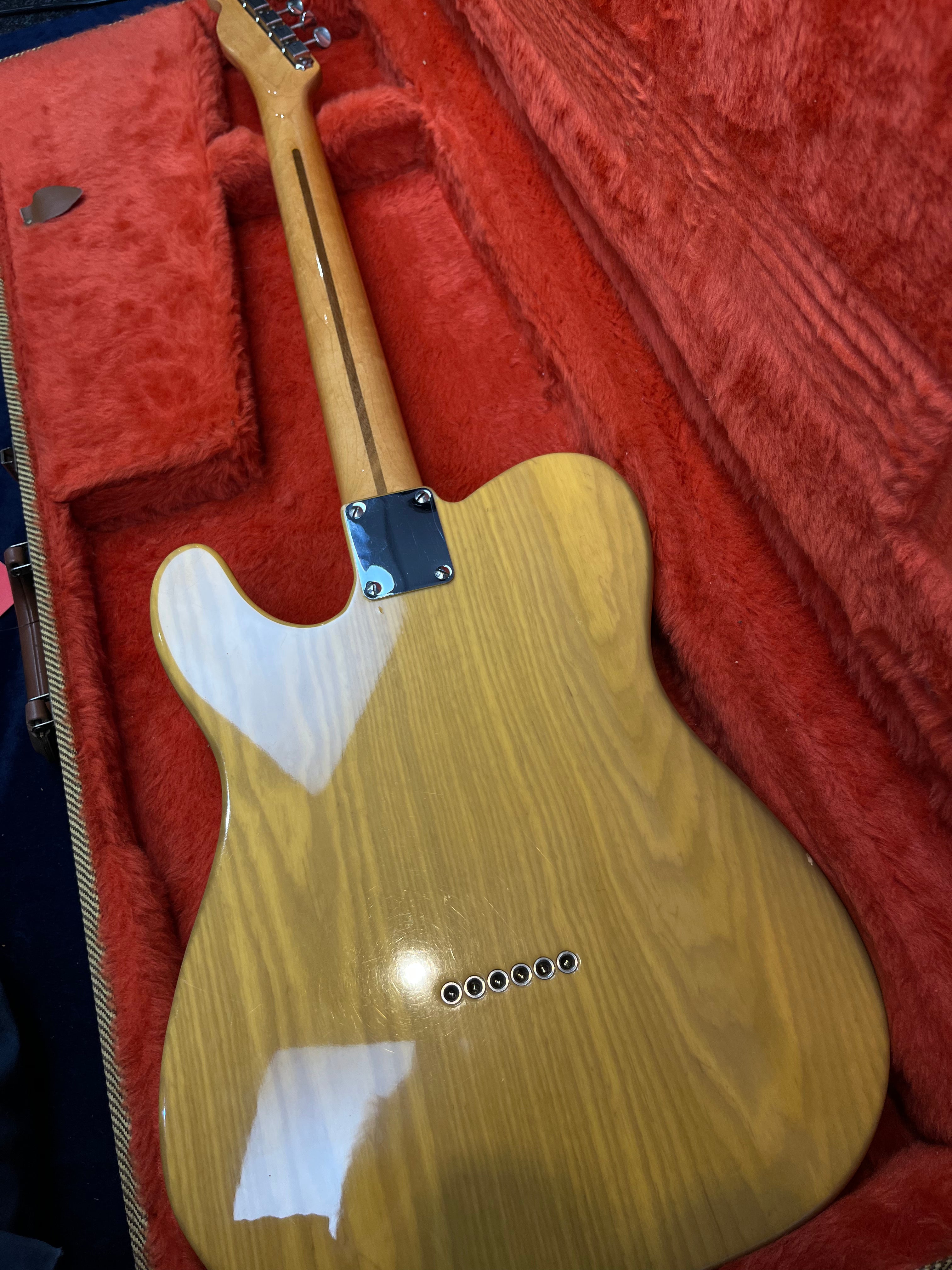 A blonde wood electric guitar rests face down in a red plush case, showing its striped grain, six bridge holes, and metal neck plate.