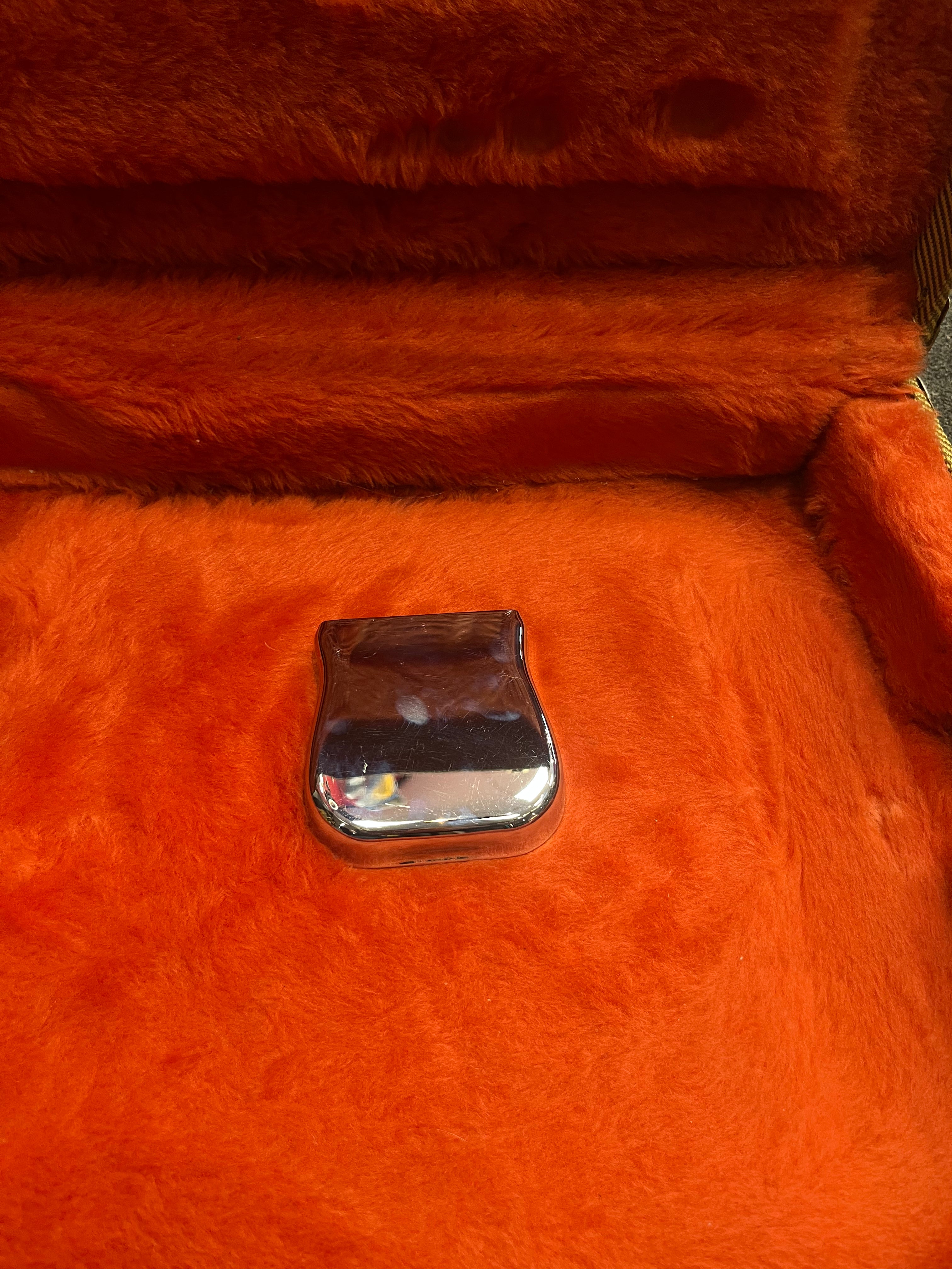 A chrome electric guitar bridge cover rests in an orange plush-lined case.