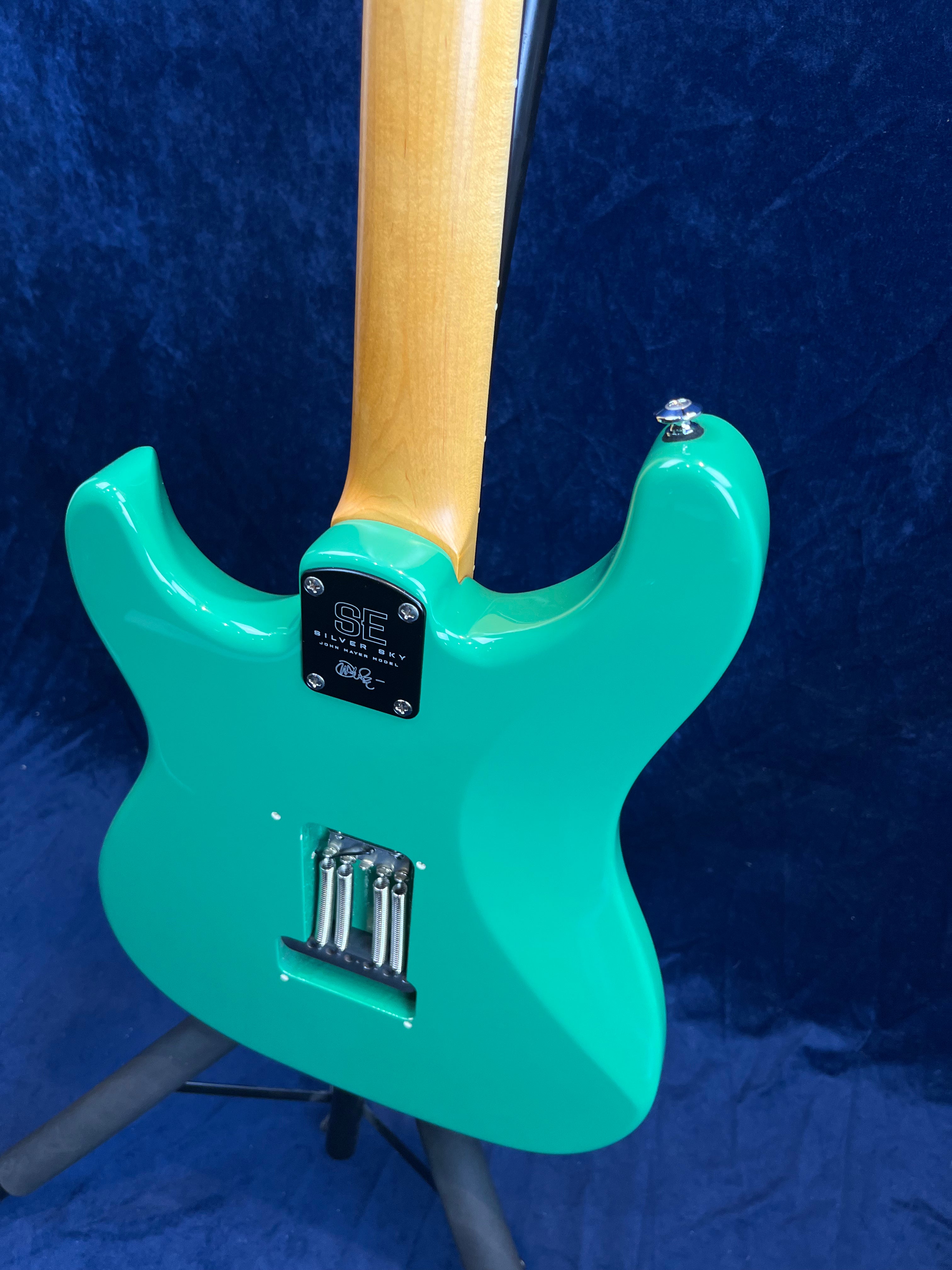 A turquoise electric guitar seen from the back, showing a natural wood neck and exposed bridge springs, set against a dark blue background.