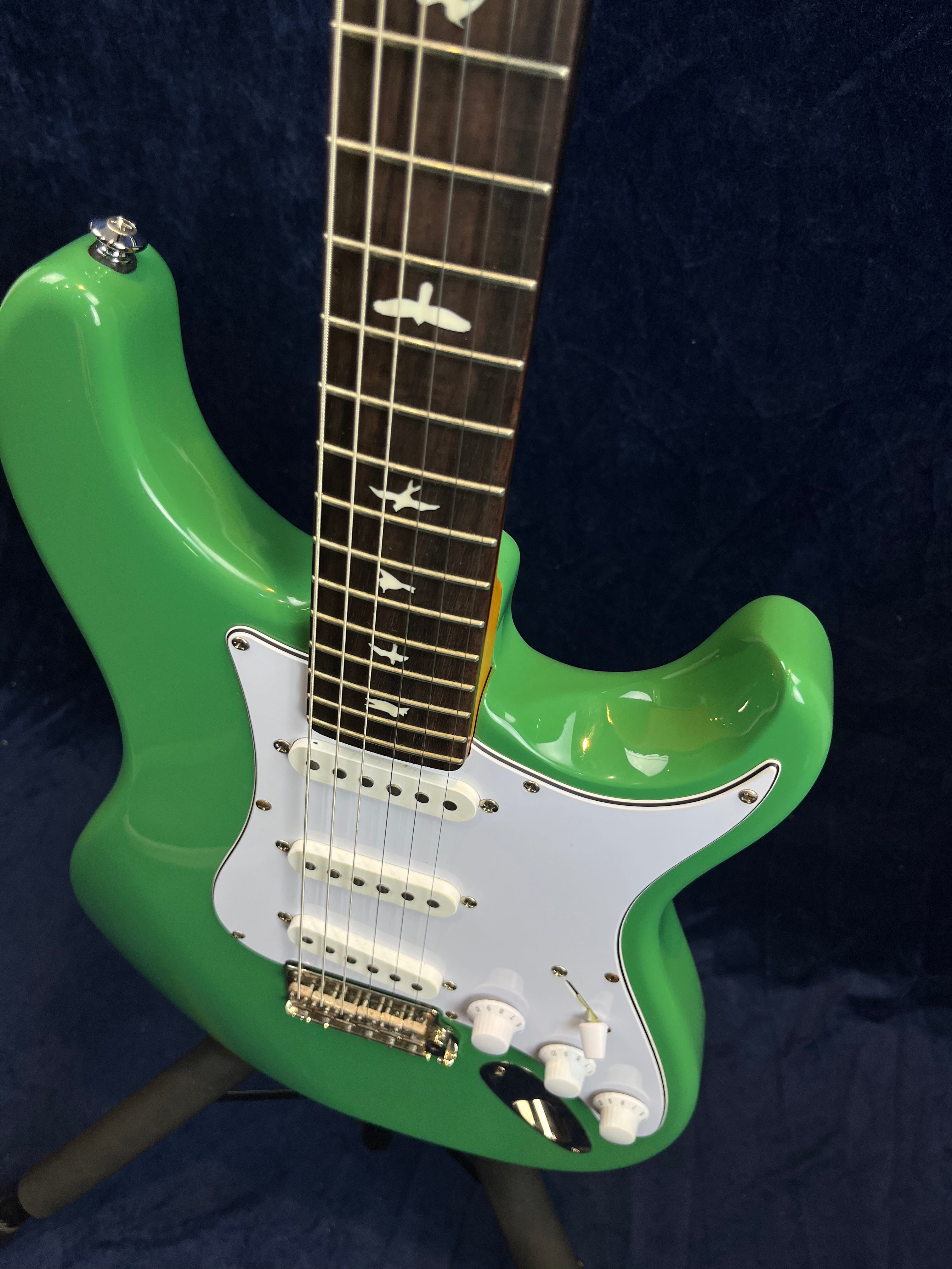 Green electric guitar with white pickguard, three single-coil pickups, three control knobs, and bird inlays on a dark fretboard.