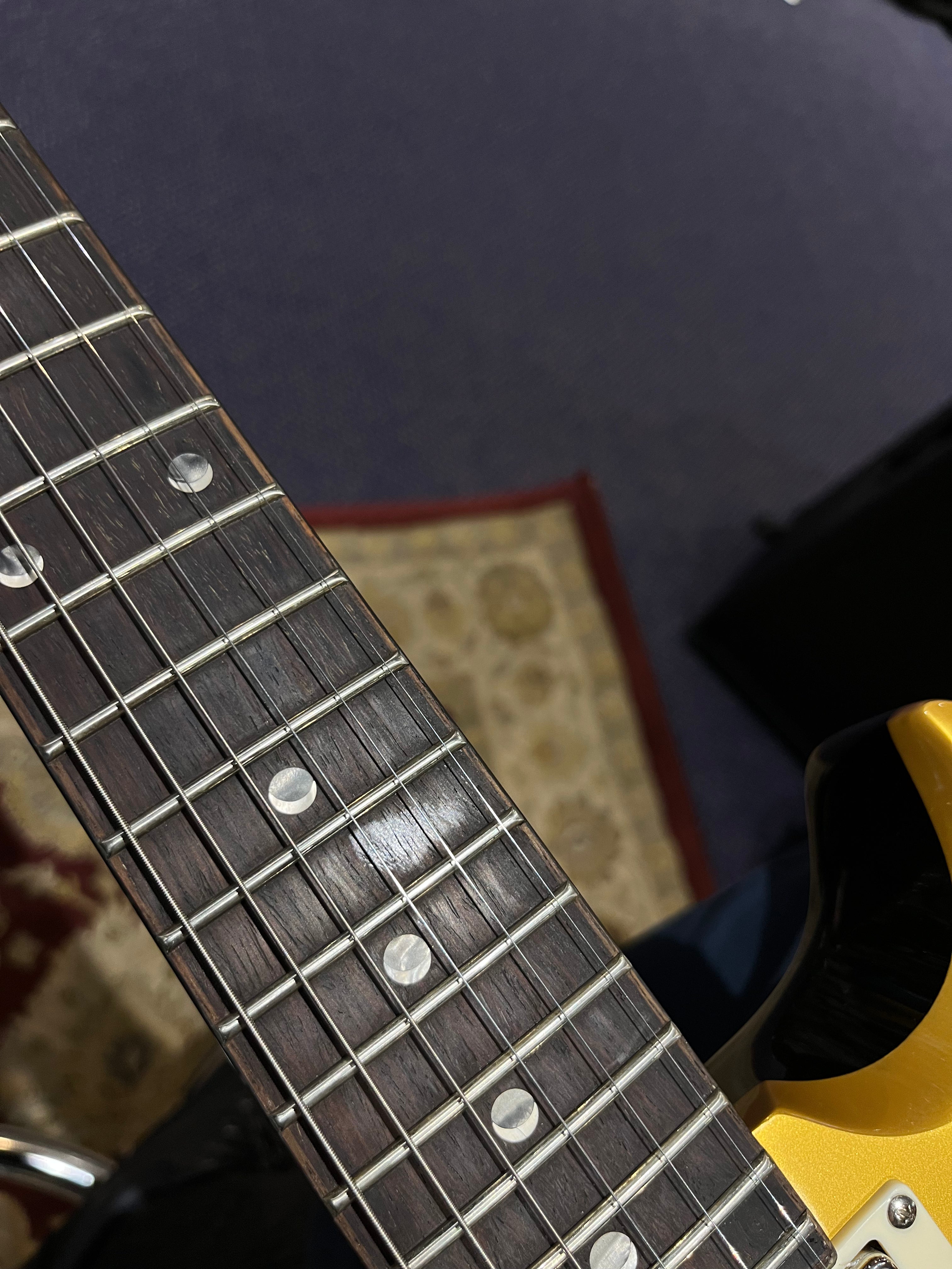 Close-up of an electric guitar’s rosewood fretboard with dot inlays, showing metal frets and part of a gold body against a patterned rug.
