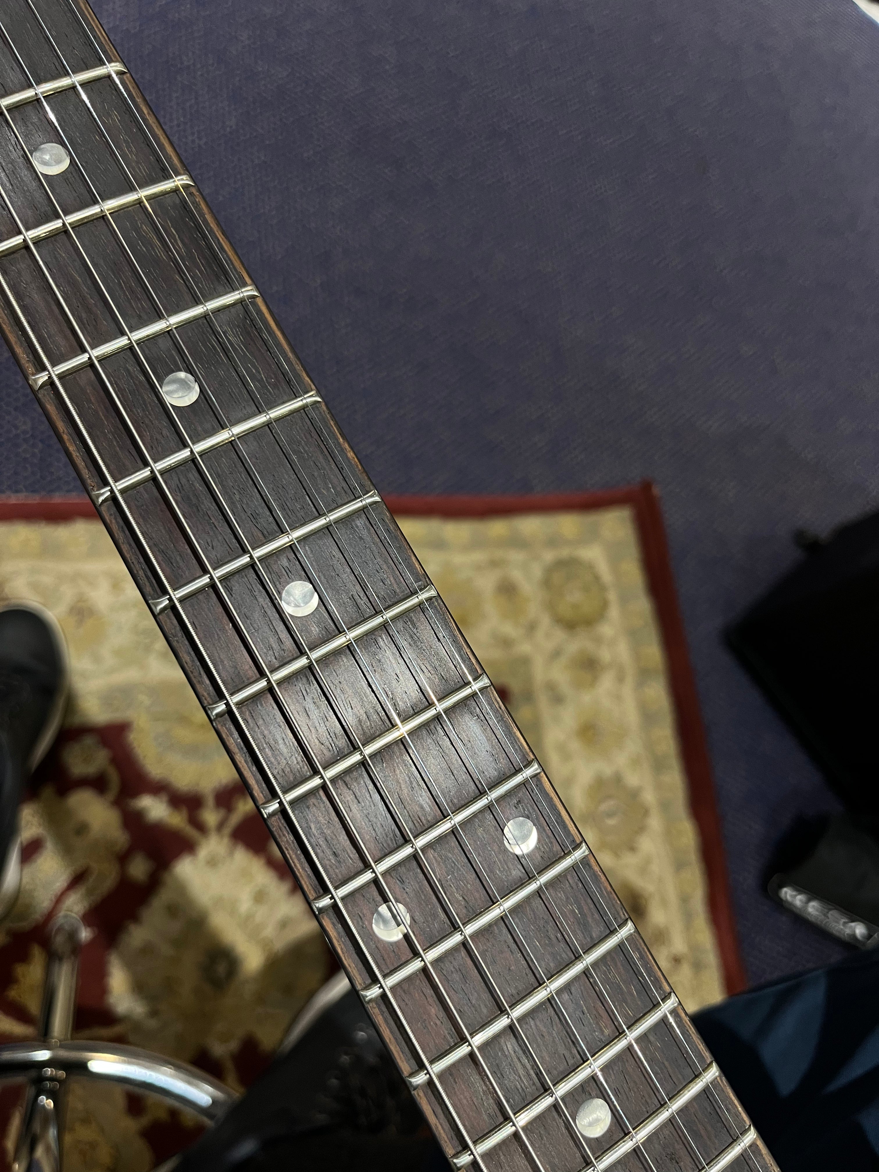 Close-up of an electric guitar neck showing frets, metal strings, and circular inlay markers; patterned rug and stool are partially visible underneath.