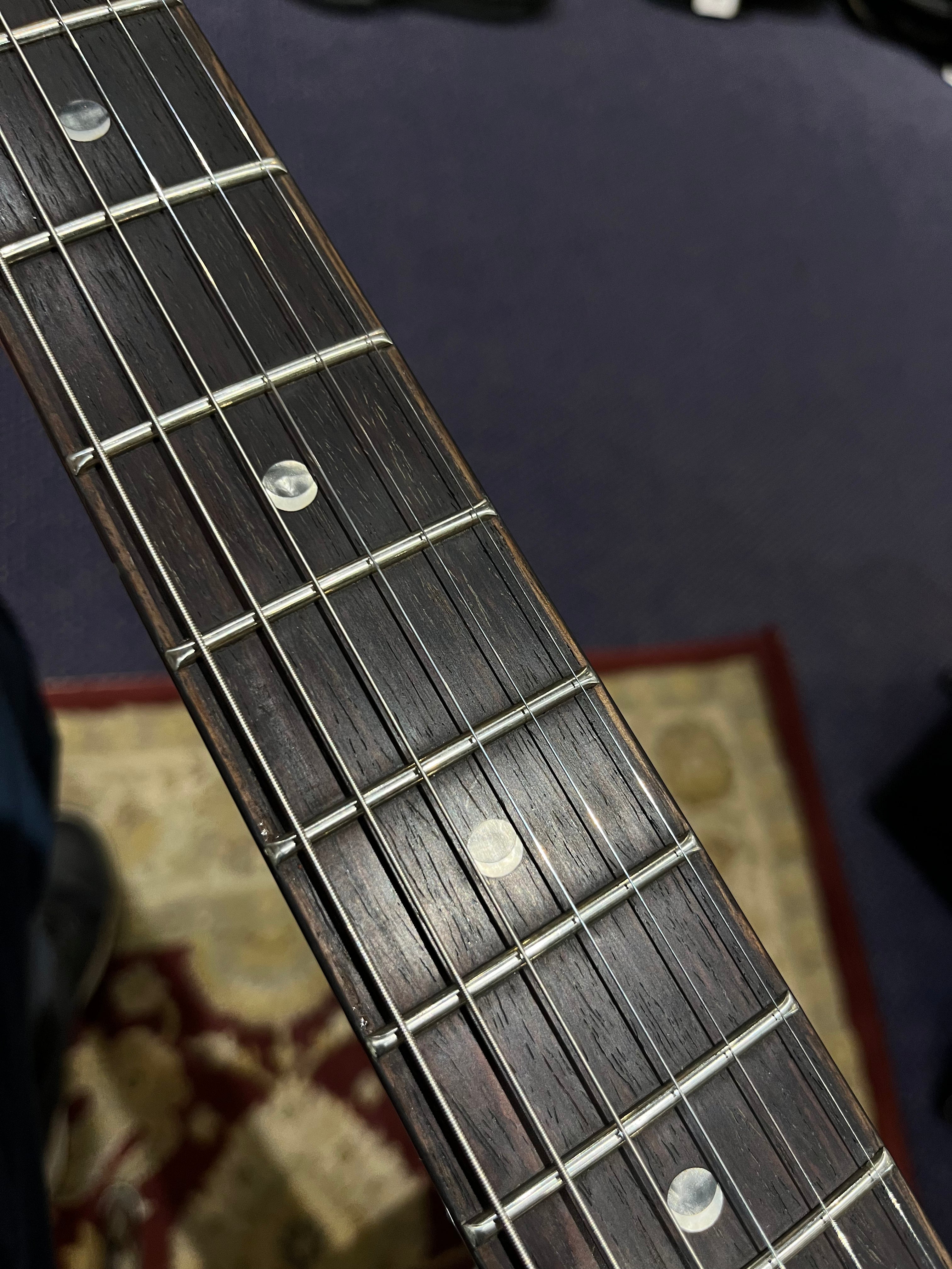 Close-up of an electric guitar fingerboard showing six metal strings, fret wires, and circular inlay markers on a dark wood background. Carpet below.