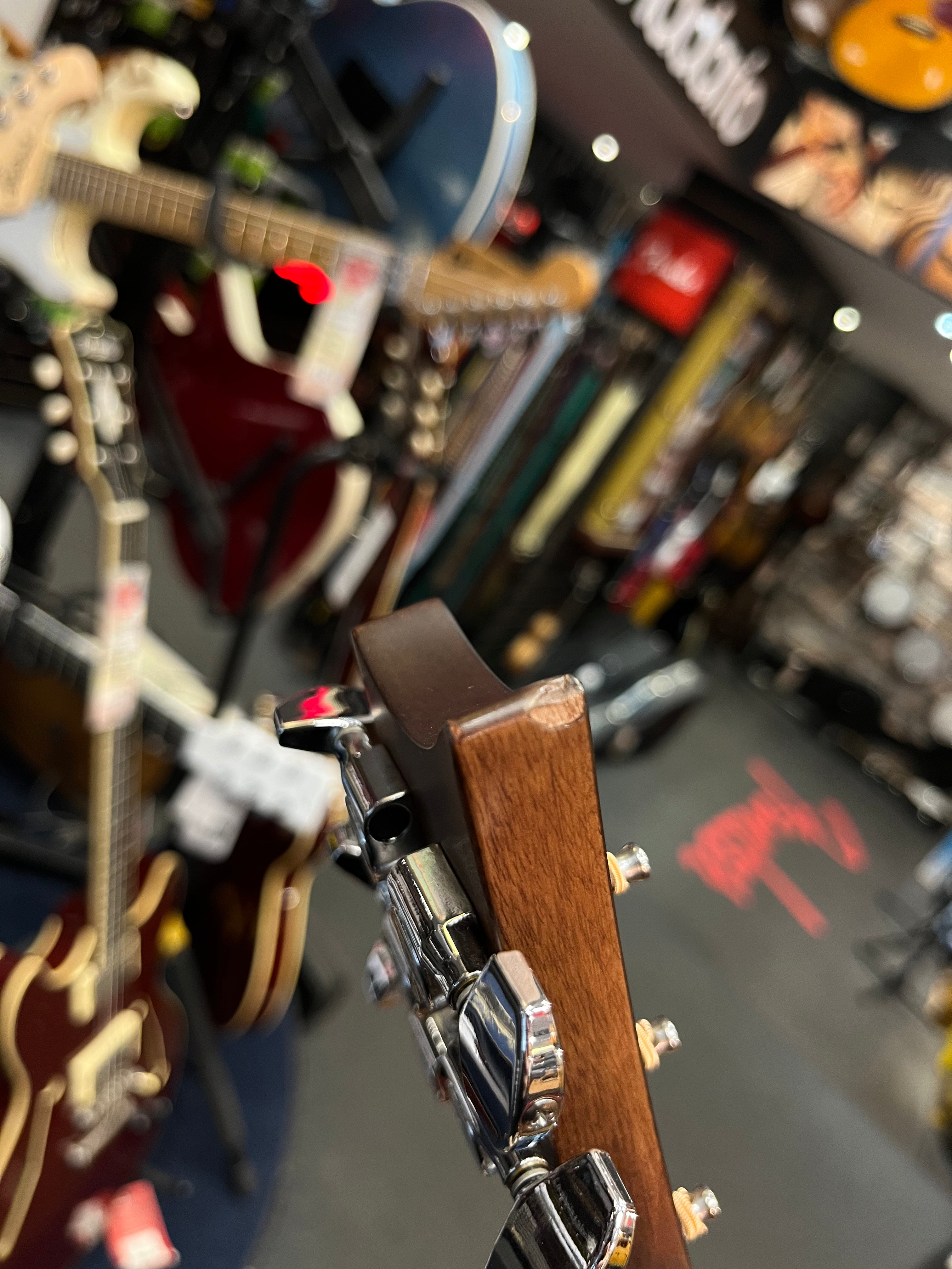 Close-up of an electric guitar headstock with chrome tuning pegs, surrounded by hanging guitars in a busy music store.