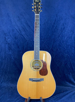 Natural wood-tone Cort acoustic guitar with a tortoiseshell pickguard, decorative rosette, and gold tuning pegs, set against a dark blue background.